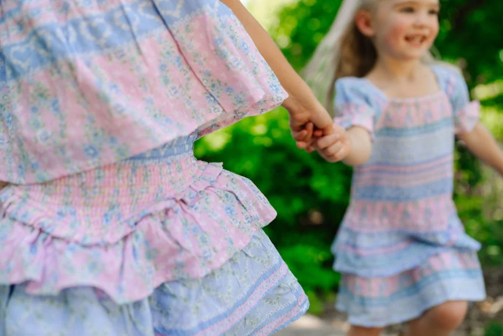 Two young girls holding hands and walking outdoors in a green, leafy park, wearing matching pastel-colored dresses with floral patterns and ruffled skirts, smiling happily.