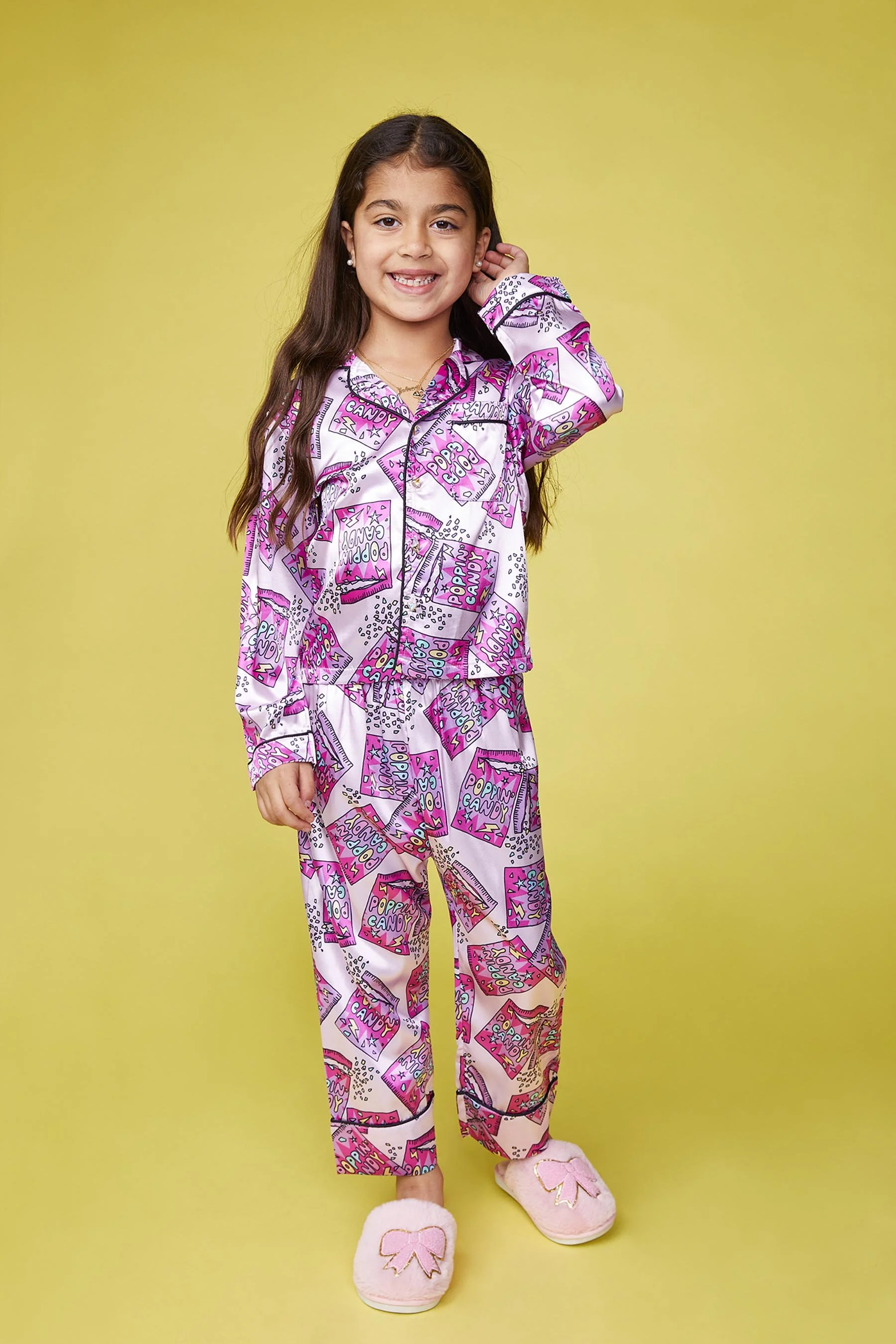 Young girl in pink and purple pop art pajamas and slippers standing in front of a yellow background, smiling and touching her hair.