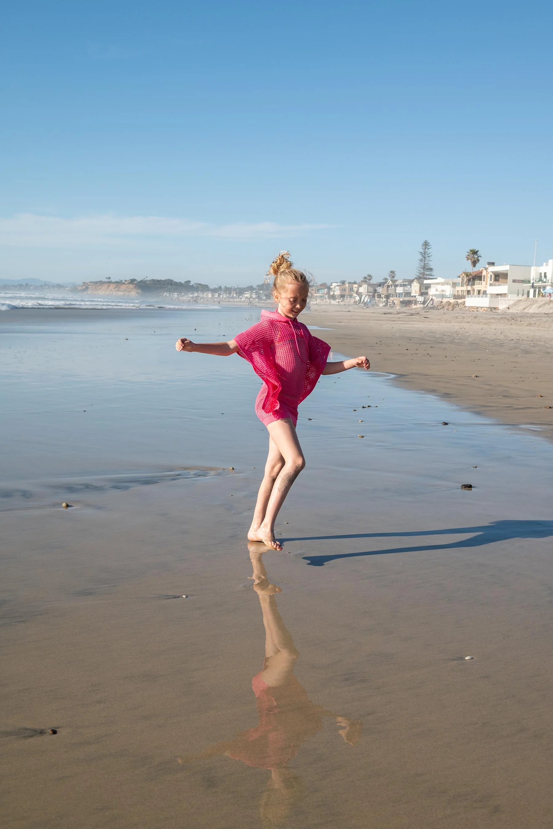 A young girl in a pink poncho playing on the wet sand at a beach during daytime, with houses and palm trees in the background, and a clear blue sky overhead.