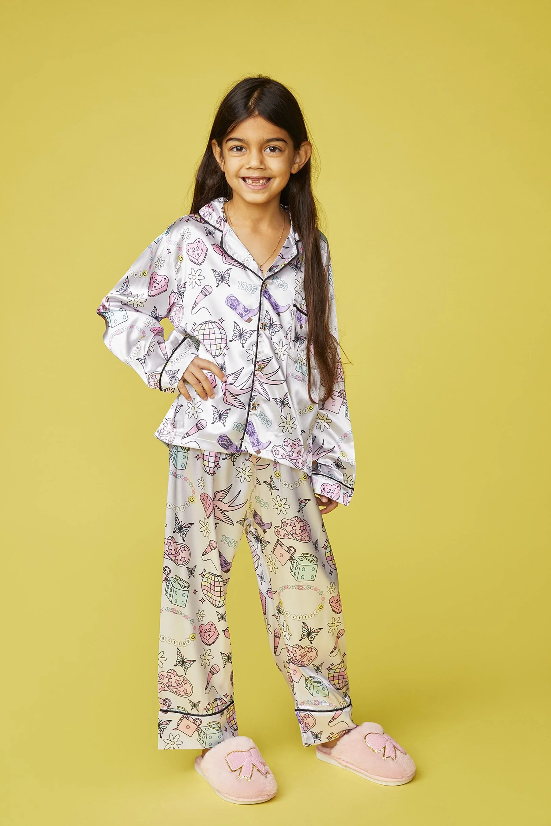 Young girl in colorful pajama set and pink slippers standing with hand on hip, smiling, against a yellow background.