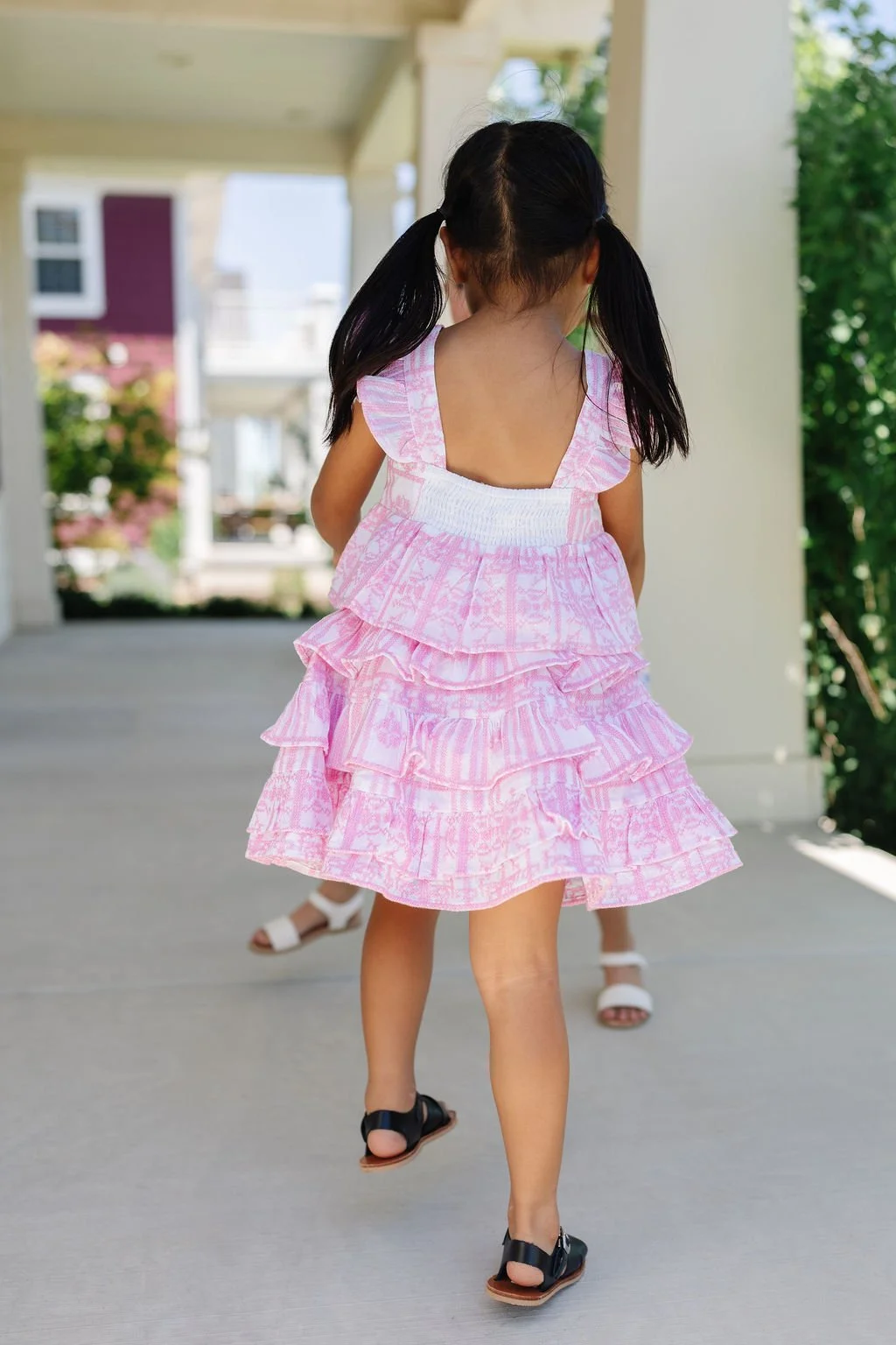 A young girl with two ponytails wearing a pink ruffled dress and black sandals walking on a porch.