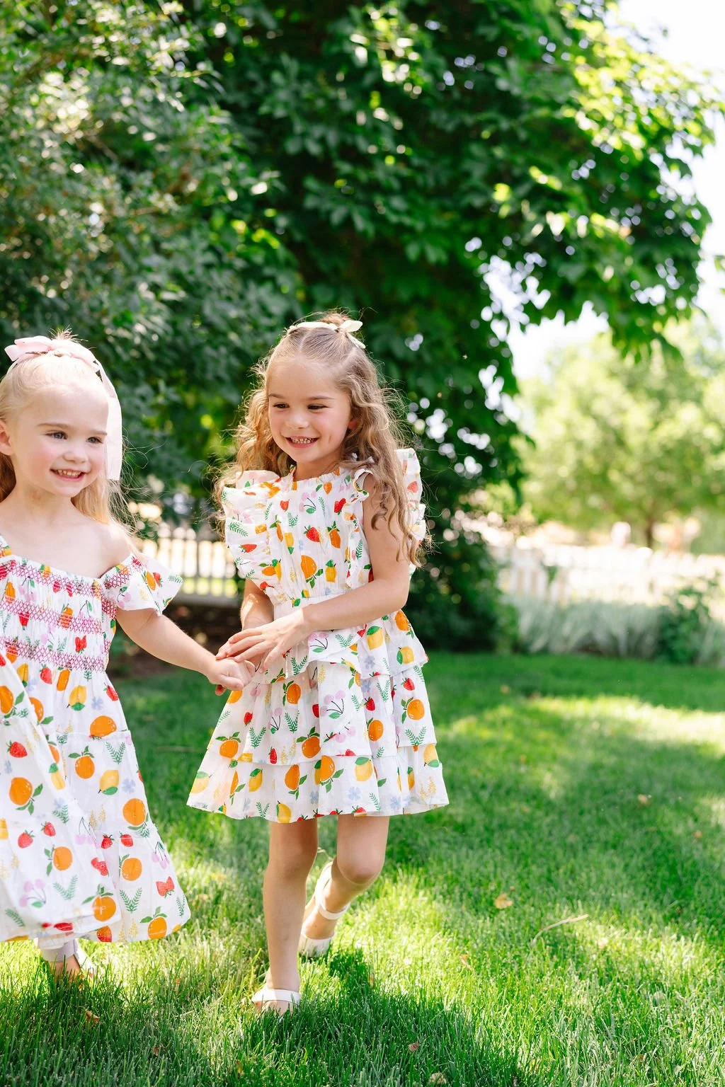 Two young girls in matching colorful fruit-patterned dresses holding hands and smiling in a lush green yard.