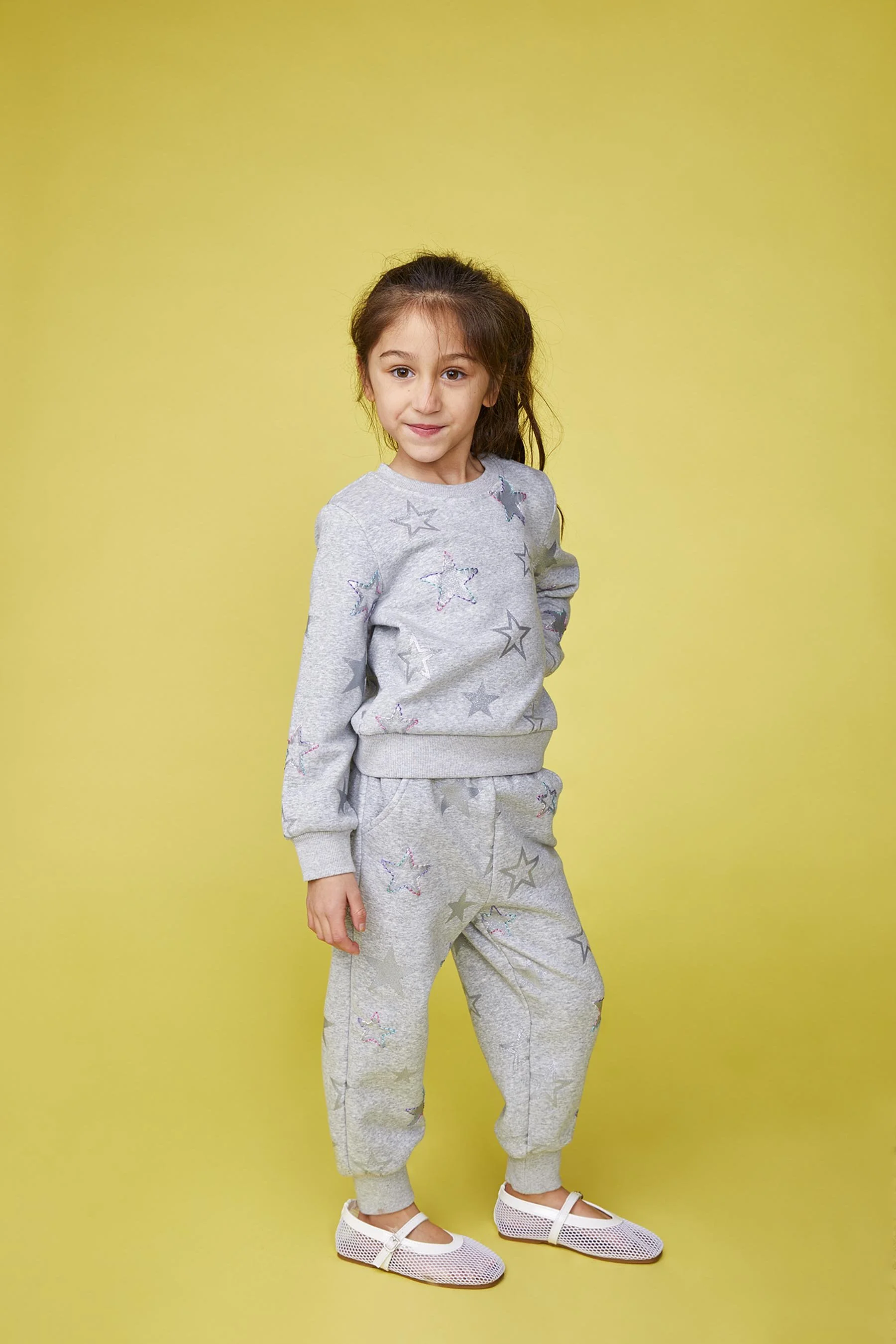 A young girl with brown hair in a ponytail wearing a gray star-patterned sweatshirt and sweatpants, white mesh shoes, standing against a yellow background.