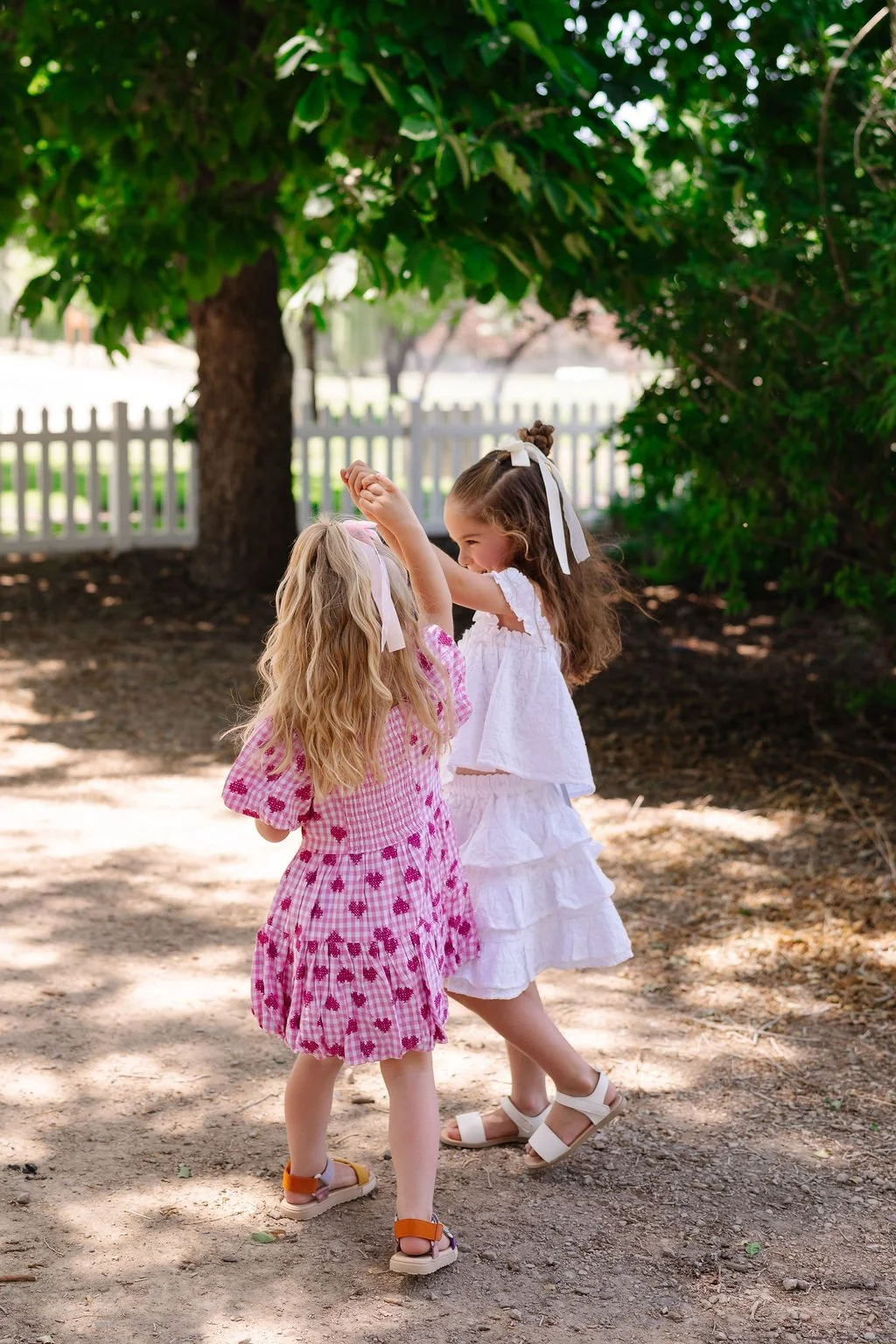 Two young girls dancing and playing together outdoors in a shaded area with trees, a wooden fence, and a grassy field in the background. One girl wears a pink checkered dress with red hearts, and the other wears a white ruffled dress. They are holdin