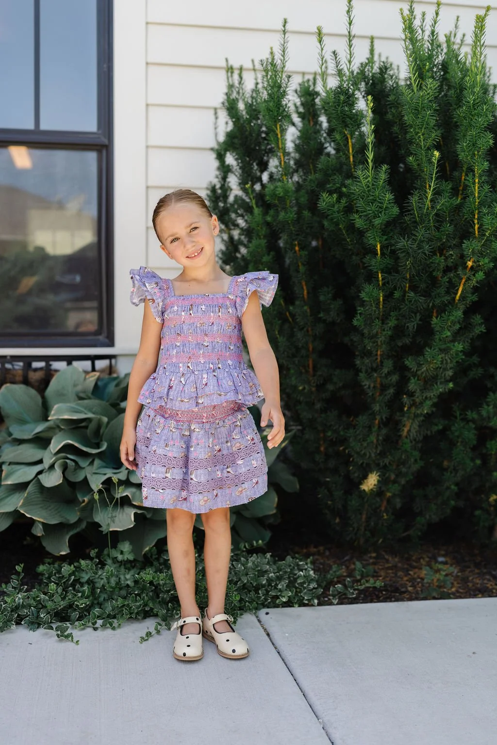 A young girl with blonde hair in a purple dress and white shoes standing outside near a house with greenery.