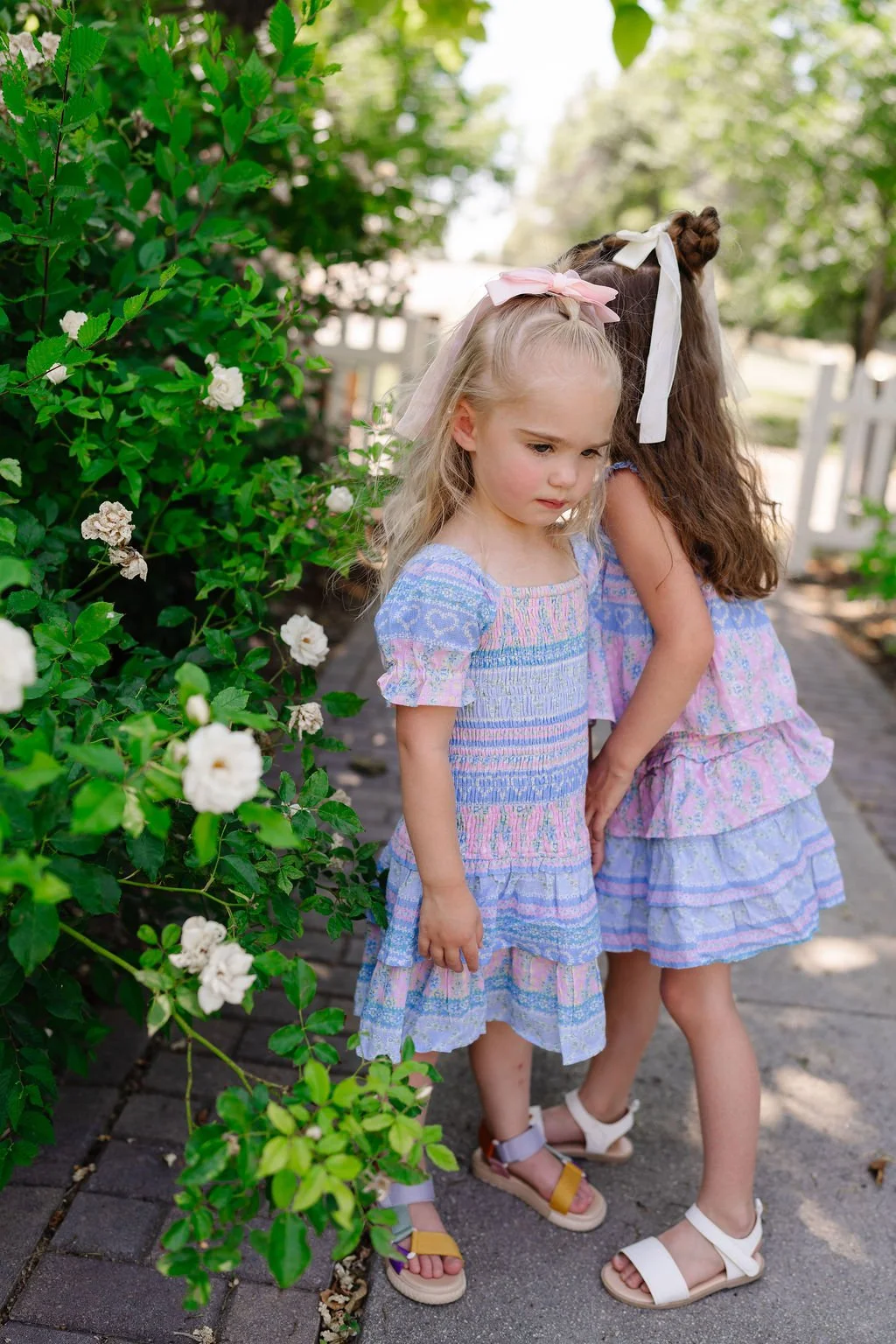 Two young girls with ribbon bows in their hair, wearing pastel-colored dresses, stand closely together outdoors on a brick path next to green bushes with white flowers, under sunlight.