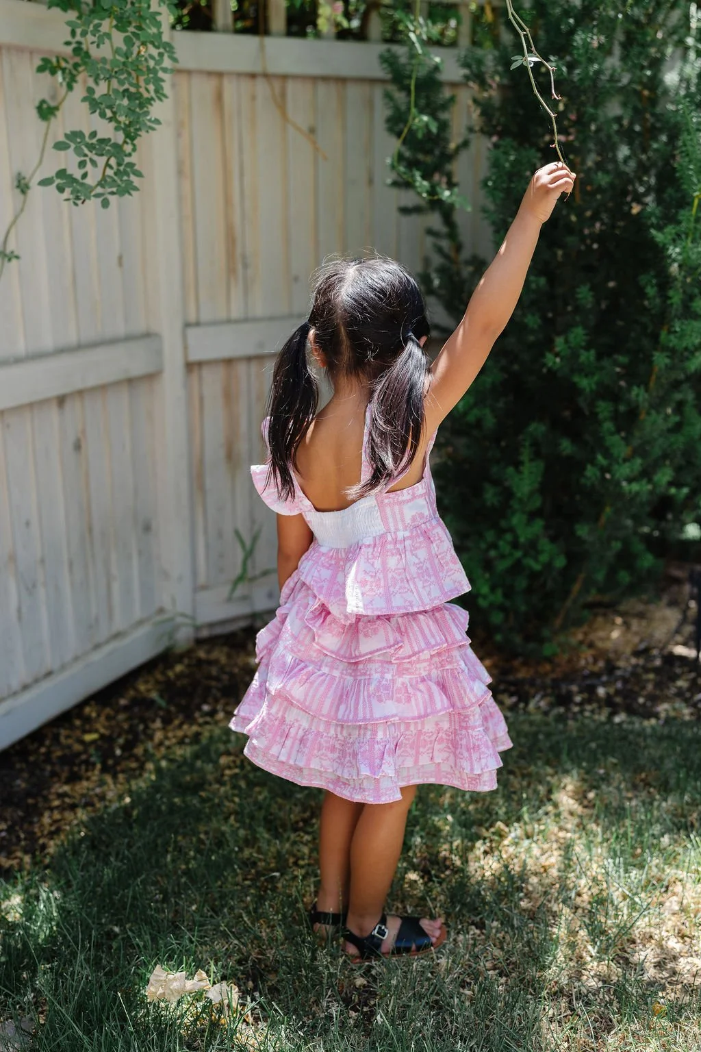 A young girl with dark hair tied in pigtails, wearing a pink ruffled dress, is raising her right arm to touch a hanging vine in a backyard with a wooden fence and green bushes.