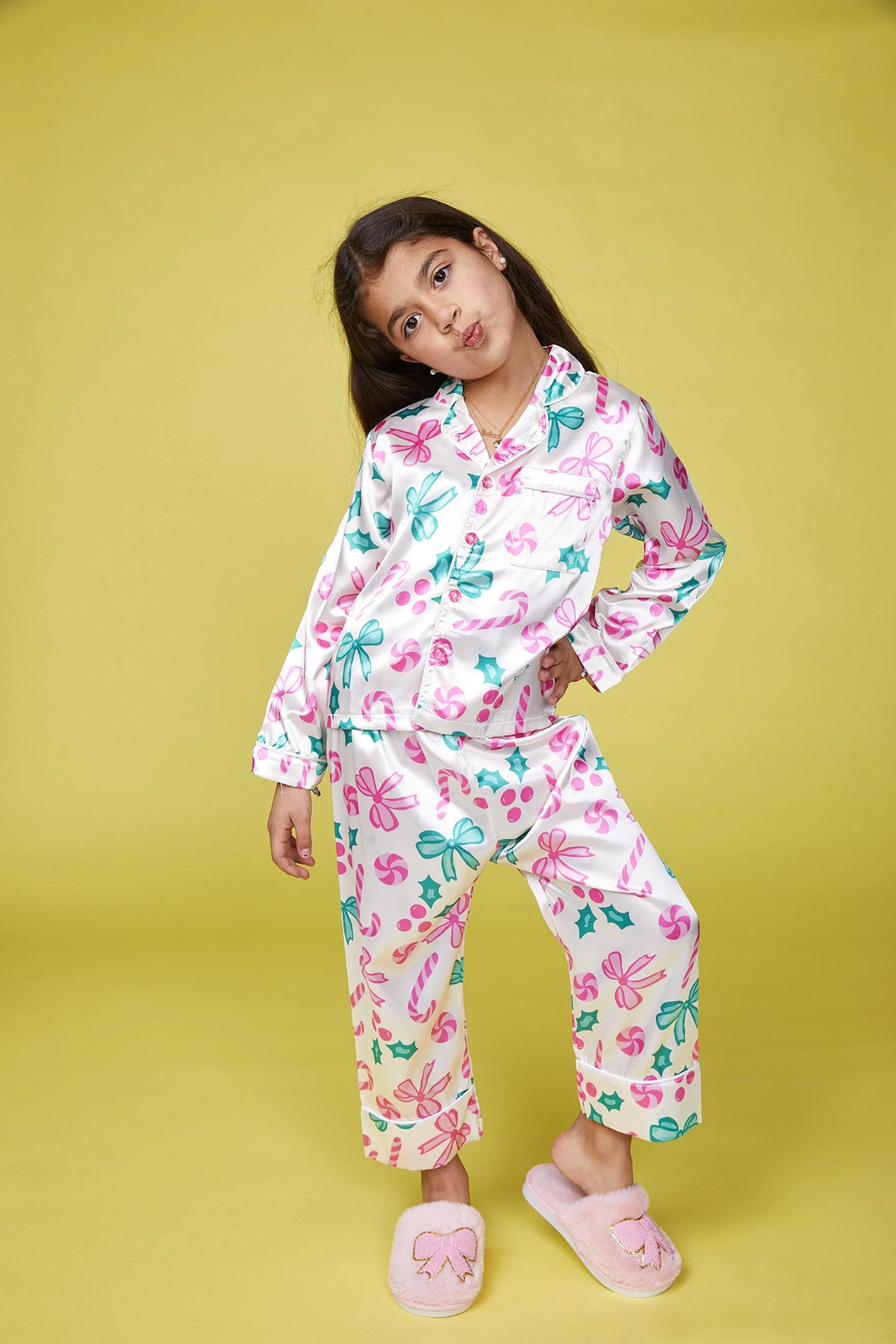 Young girl wearing Christmas-themed pajamas with pink, green, and blue candy canes, holly, and bows, standing against a yellow background, making a playful pout.