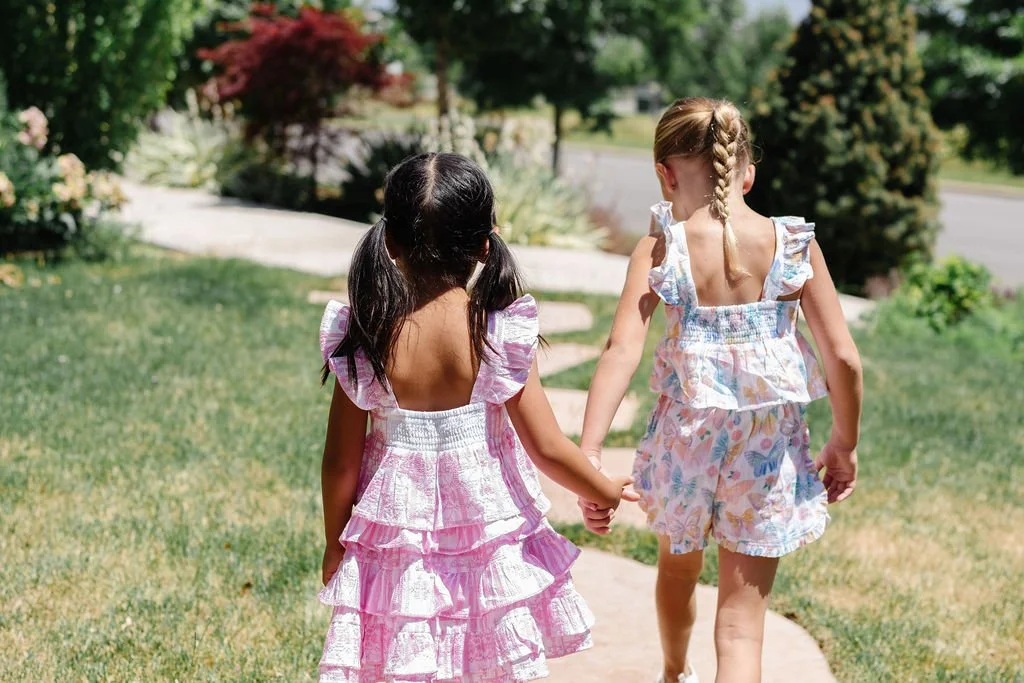 Two young girls holding hands and walking on a sidewalk in a garden or park area, wearing colorful summer dresses.
