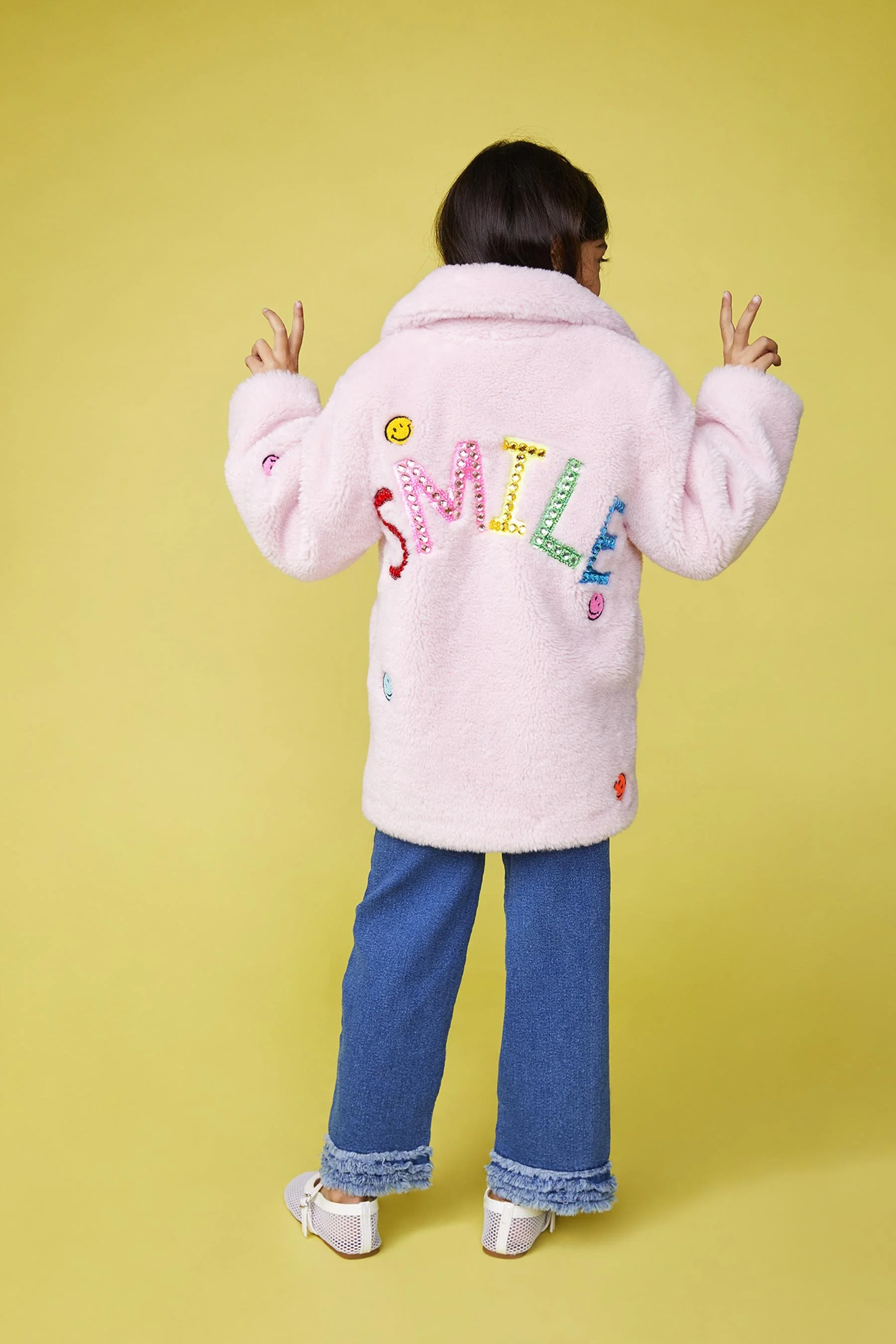 Back view of a child wearing a pink fuzzy coat with colorful, embroidered letters spelling 'SMILE' and small expressive patches, paired with wide-leg blue jeans with frayed cuffs, standing against a yellow background, making peace signs with both han