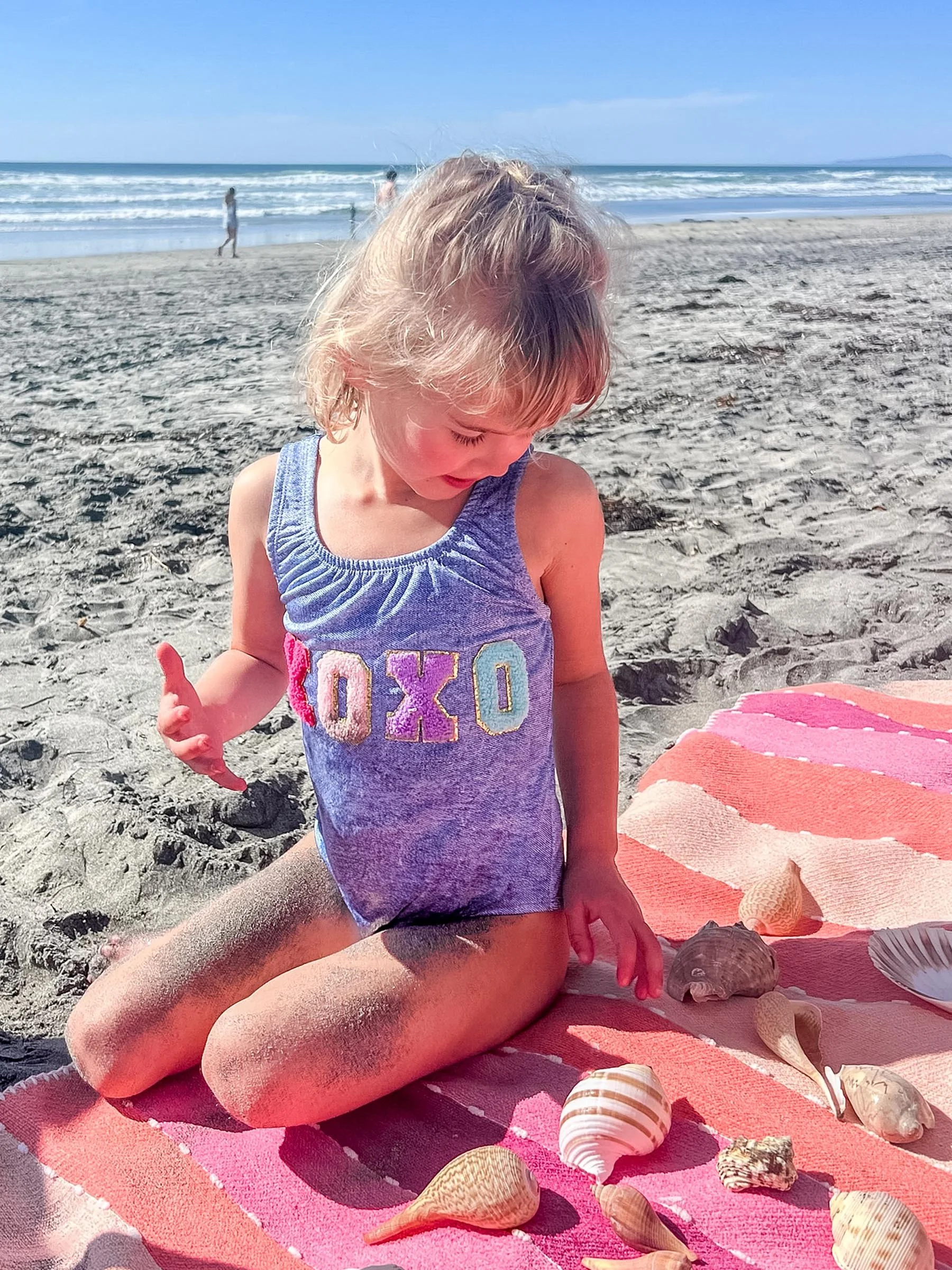 A young girl kneeling on a pink striped towel on a sandy beach, surrounded by seashells, with the ocean and a blue sky in the background.