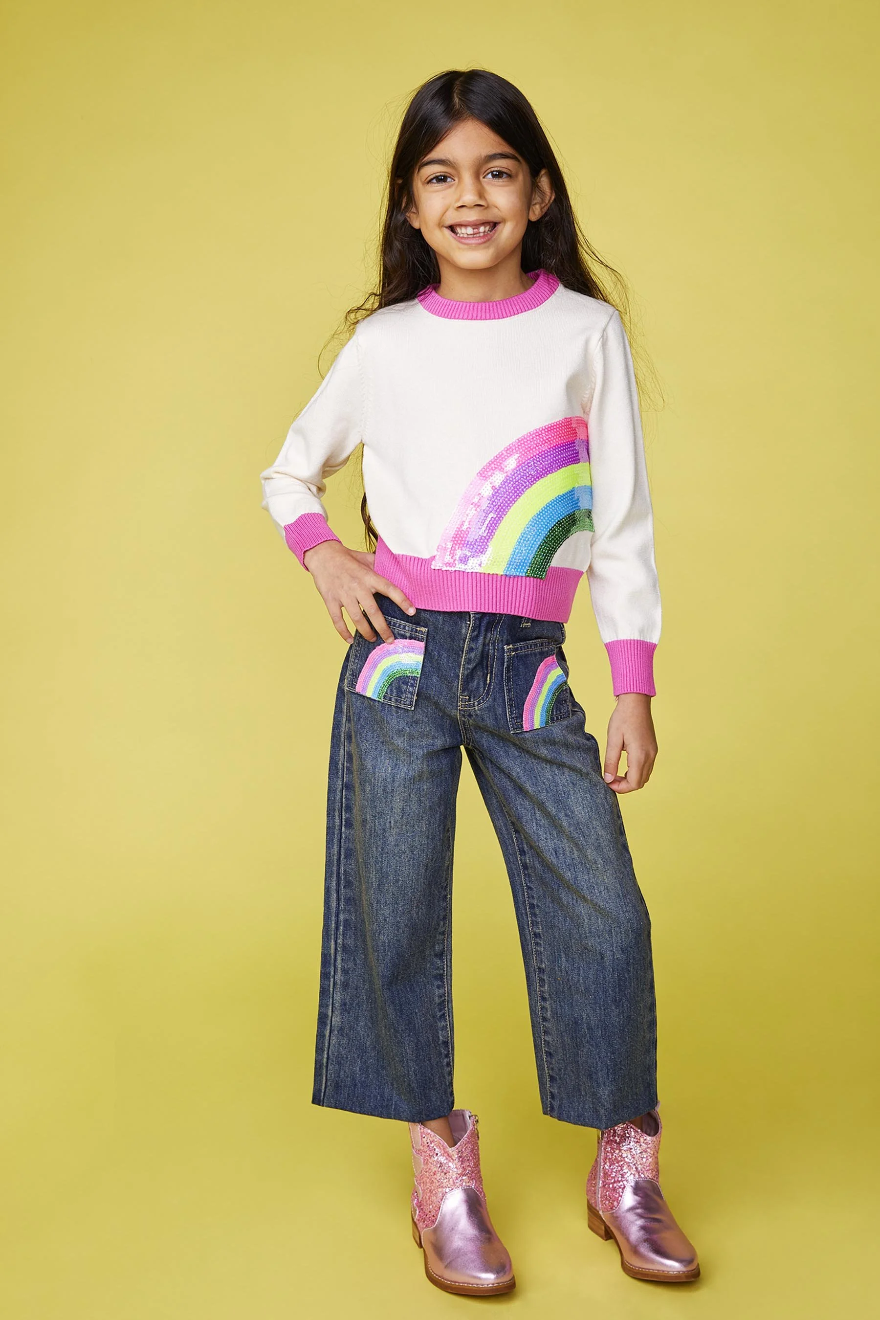 A smiling young girl with long dark hair wearing a white sweater with pink cuffs and a rainbow graphic, loose-fit jeans with rainbow patches on the pockets, and pink shiny cowboy boots, standing against a yellow background.