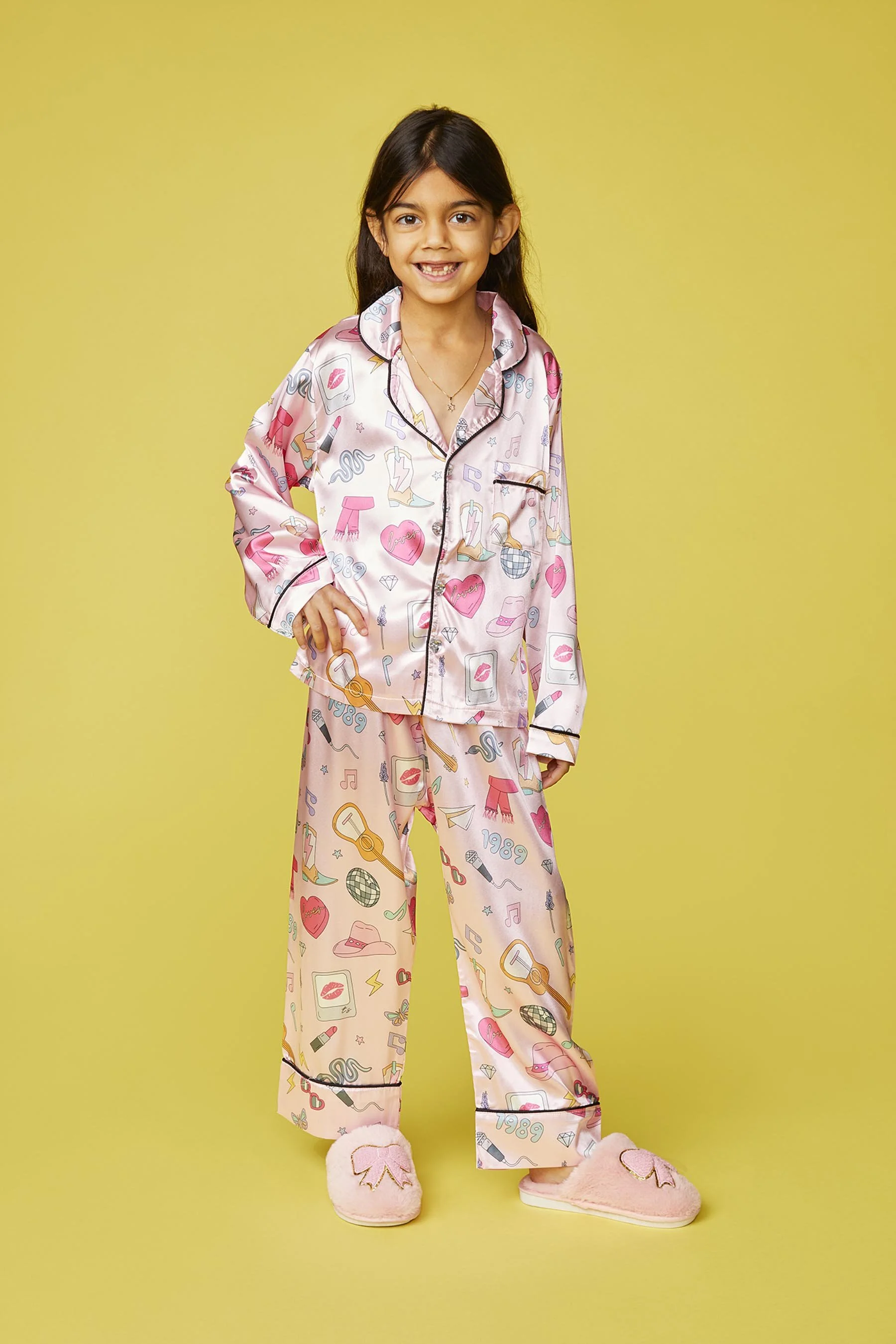 Young girl wearing pink pajama set with various colorful patterns and slippers, standing against a yellow background.