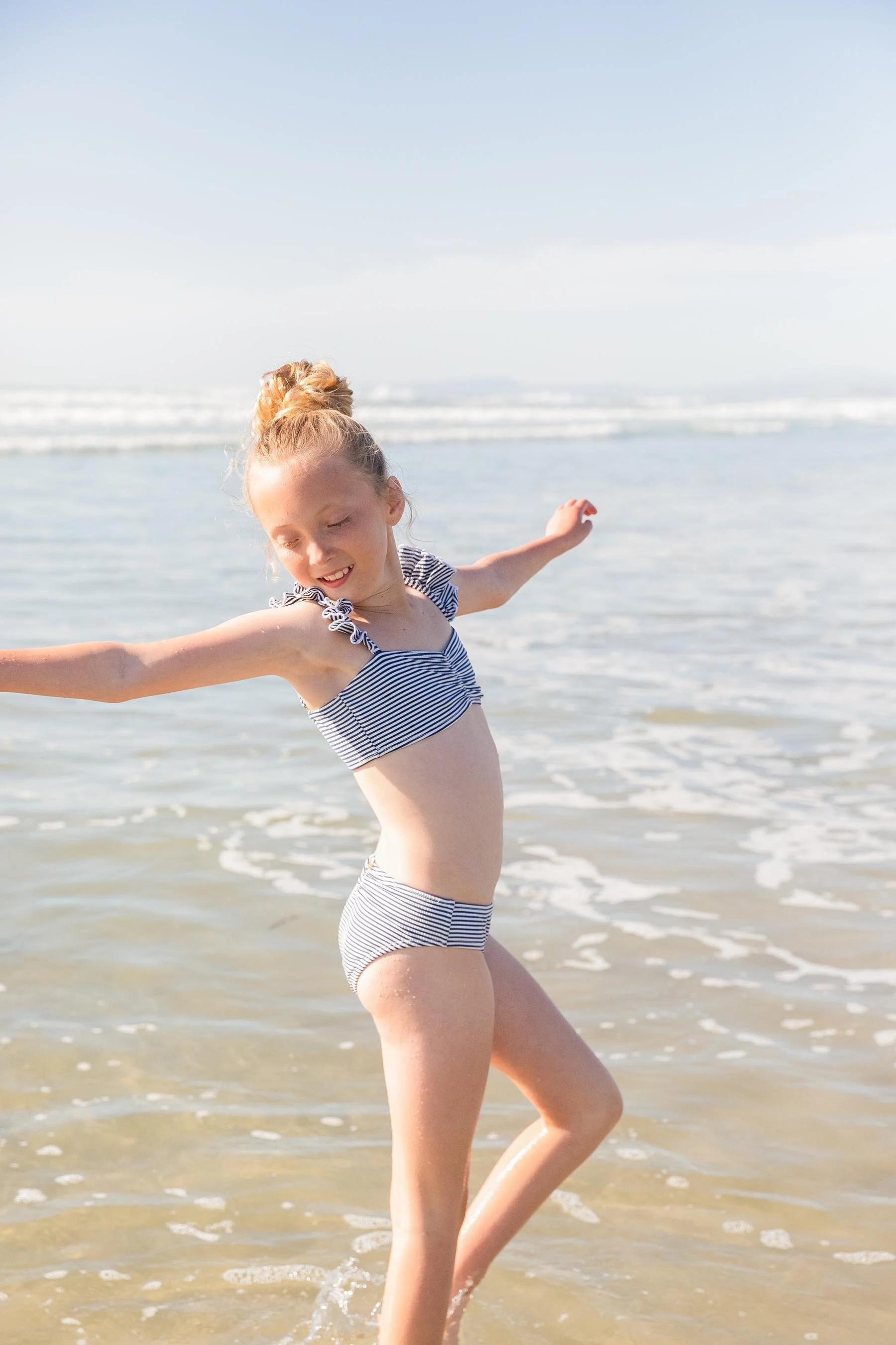 A young girl in a striped swimsuit playing and smiling in the shallow water at the beach.