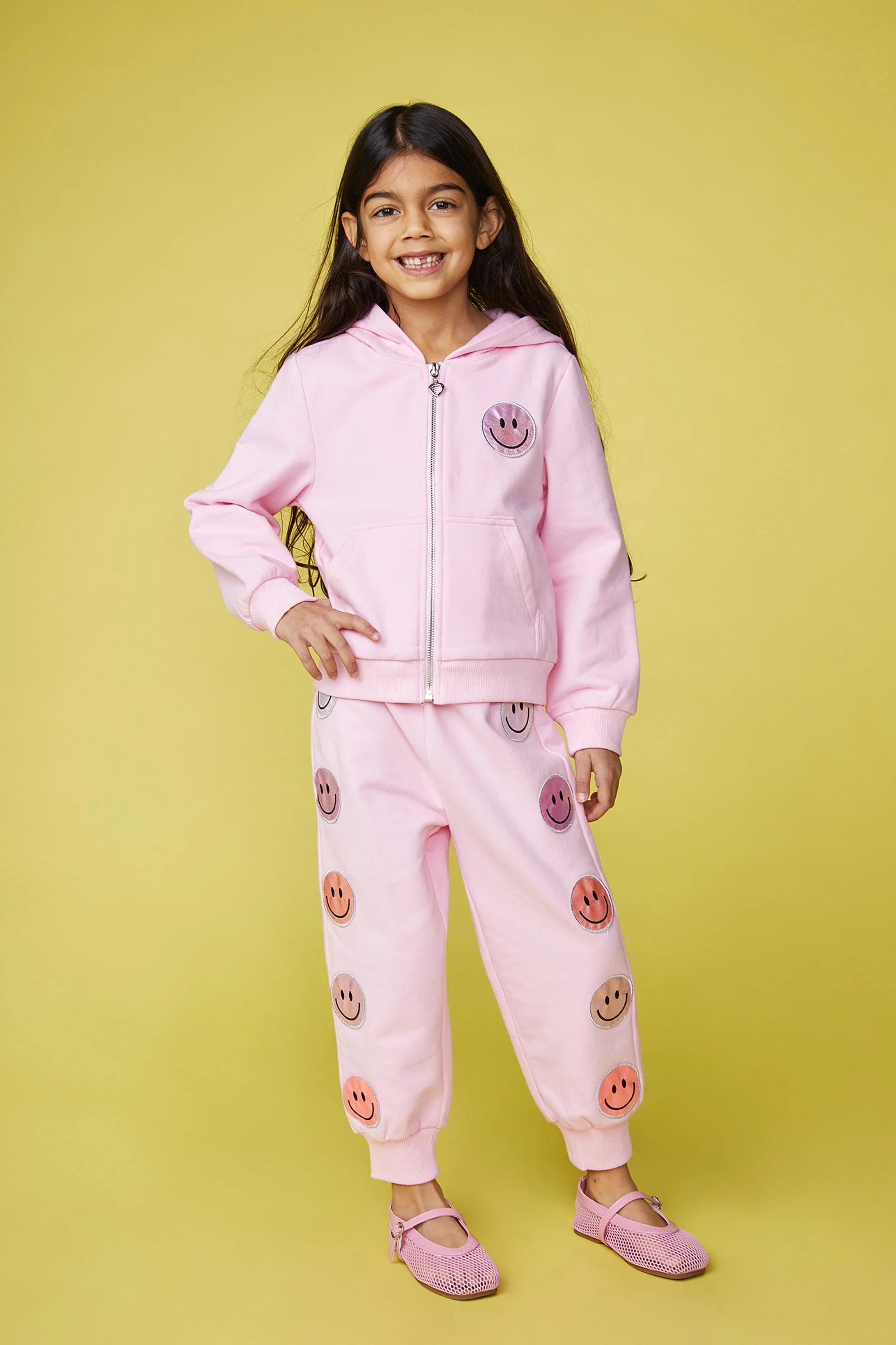 A young girl wearing a pink hoodie and matching pants with smiley faces, standing against a yellow background and smiling at the camera.