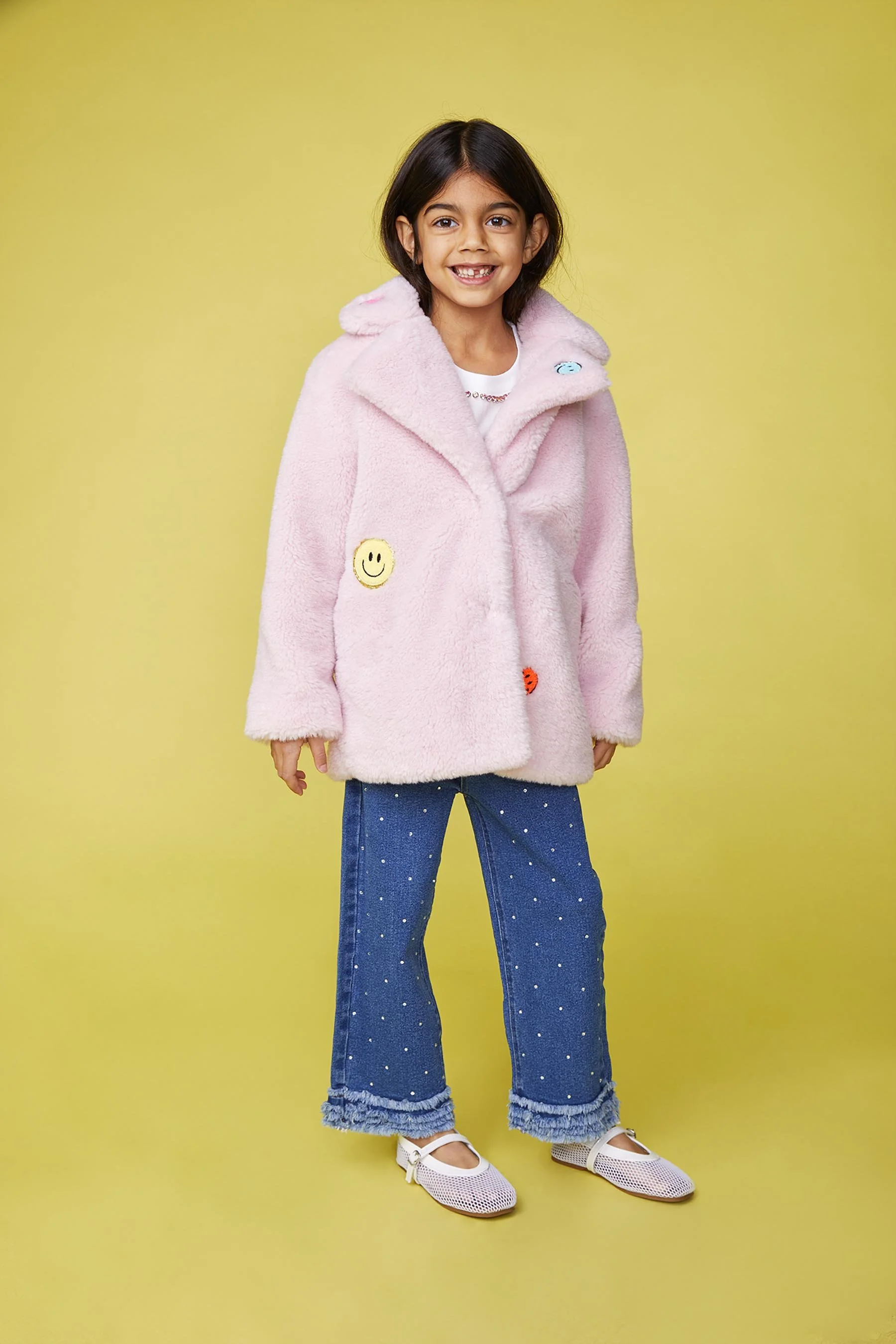 A young girl with dark hair, wearing a pink fuzzy coat with smiley face patches, blue jeans with white polka dots, and white shoes, standing against a yellow background and smiling.