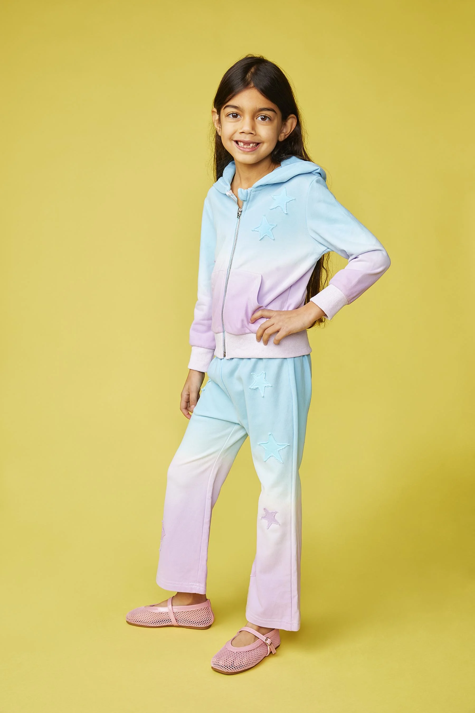 A young girl with long dark hair wearing a pastel rainbow-colored hoodie and matching pants with star patterns, pink shoes, standing against a yellow background.