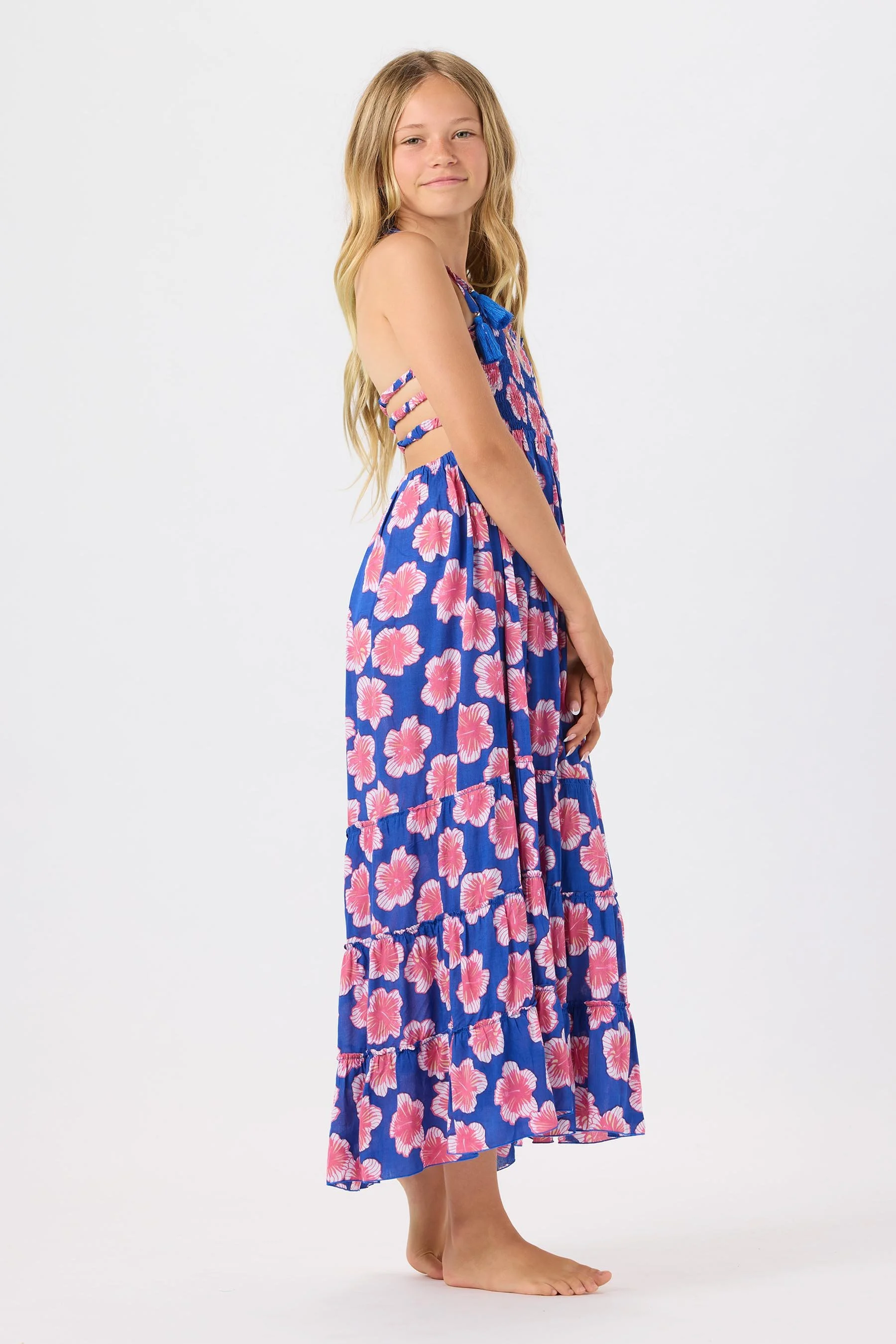 Young girl with long blonde hair wearing a blue floral maxi dress with pink flowers, standing barefoot against a white background.