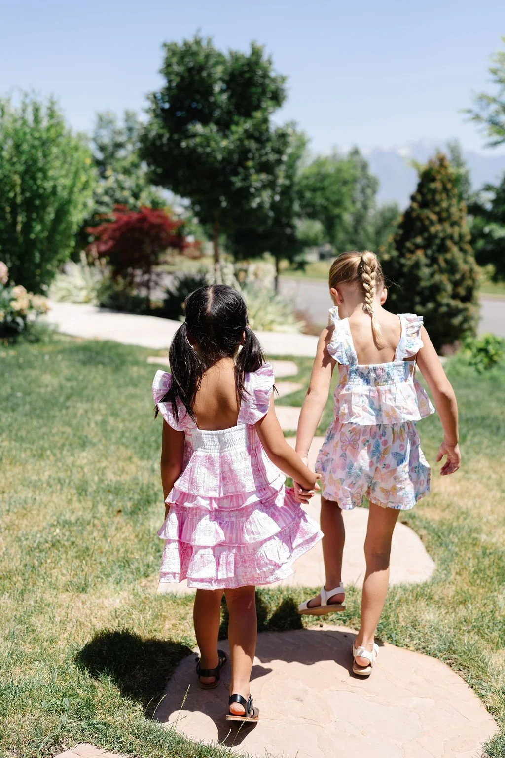 Two young girls walking hand in hand on a sunny day in a garden with trees and bushes, dressed in colorful summer outfits.