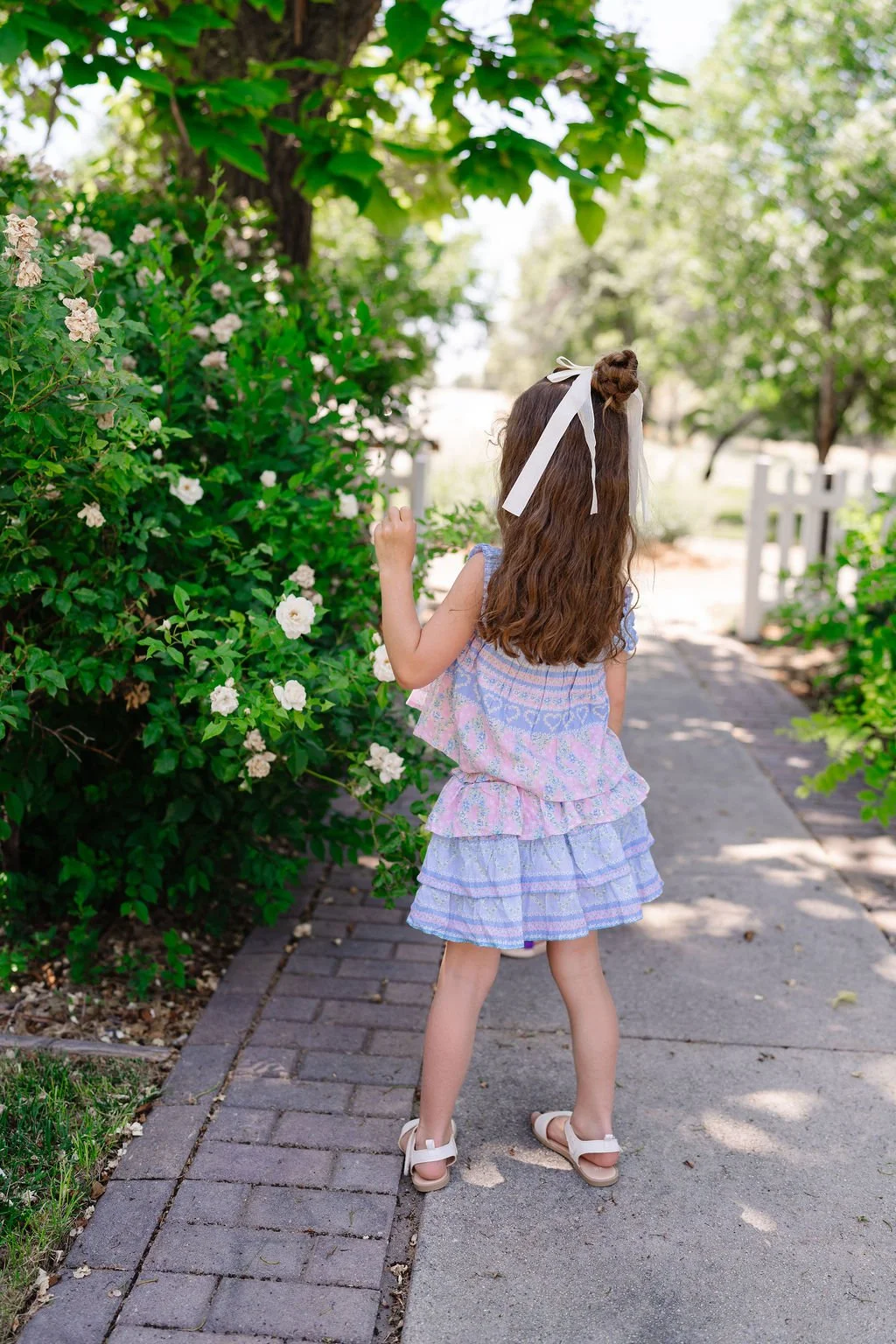 A young girl with long brown hair wearing a pastel-colored ruffled dress and white sandals is standing outdoors on a sidewalk, reaching out to touch white flowers on a bush. She has a white ribbon in her hair and is facing away from the camera.