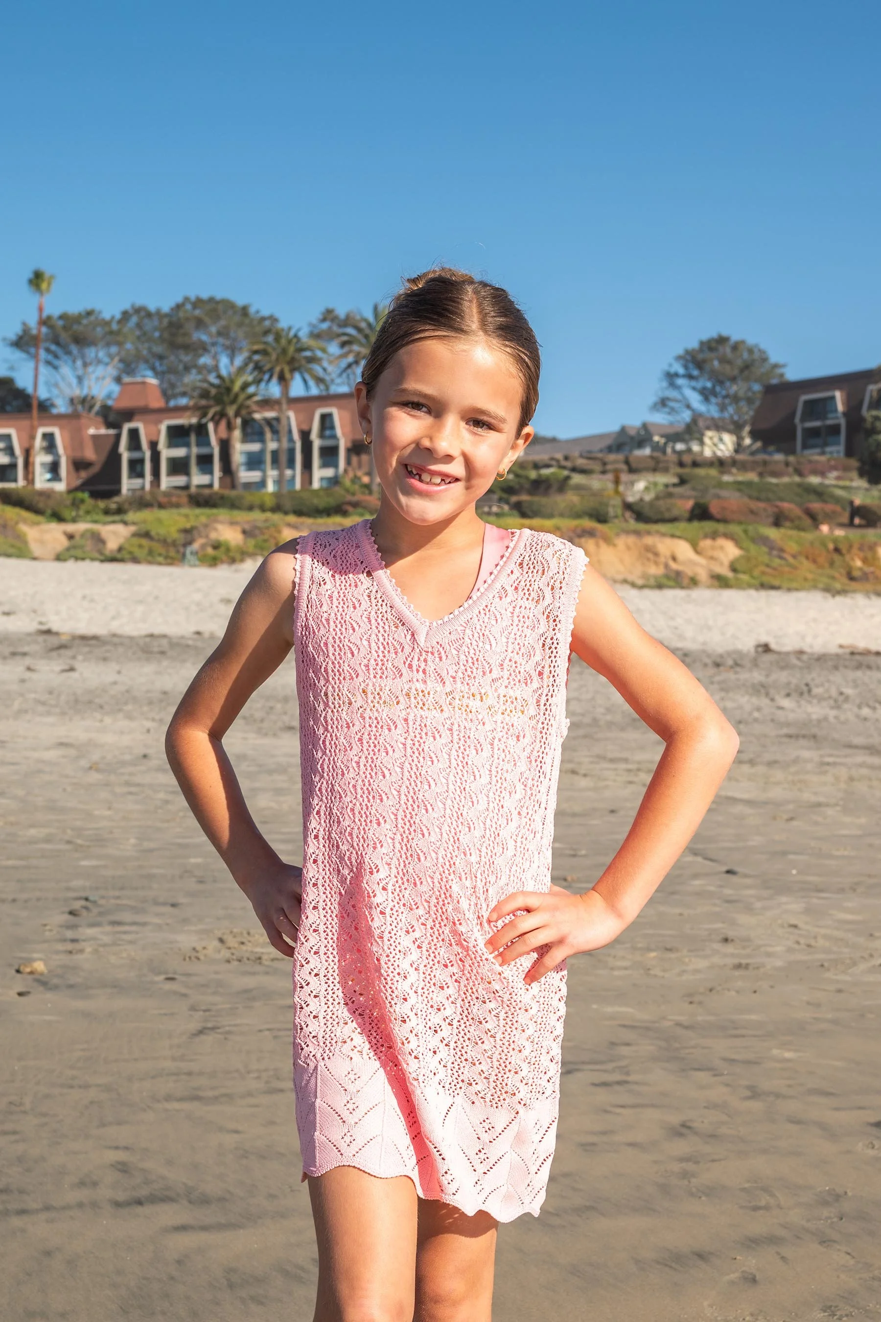 A young girl in a pink crochet dress standing on a beach with her hands on her hips, smiling at the camera. In the background, there are houses with palm trees and a clear blue sky.