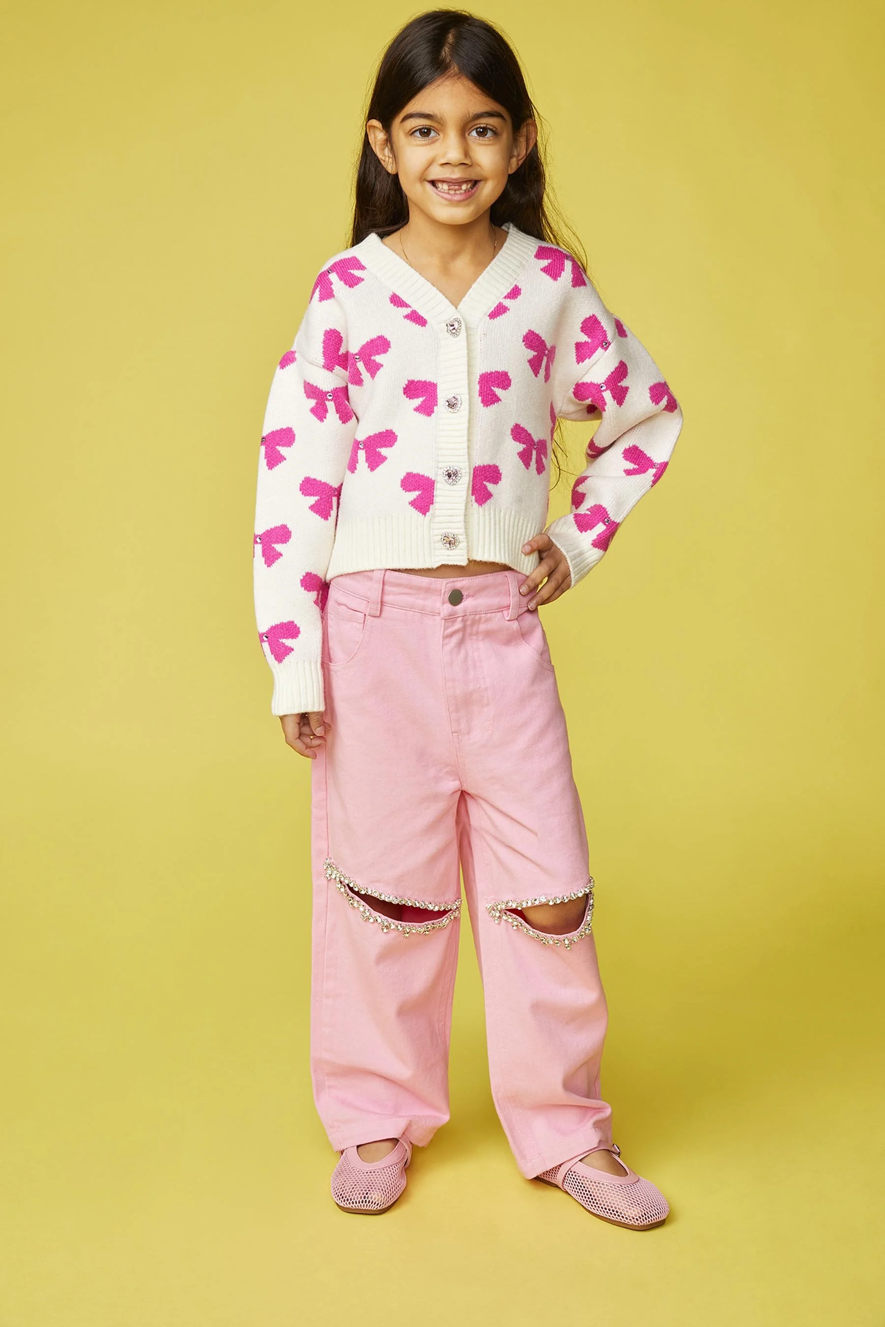 A young girl with long dark hair, smiling, wearing a white sweater with pink bow patterns, pink ripped pants with rhinestone chains, and pink shoes, standing against a yellow background.