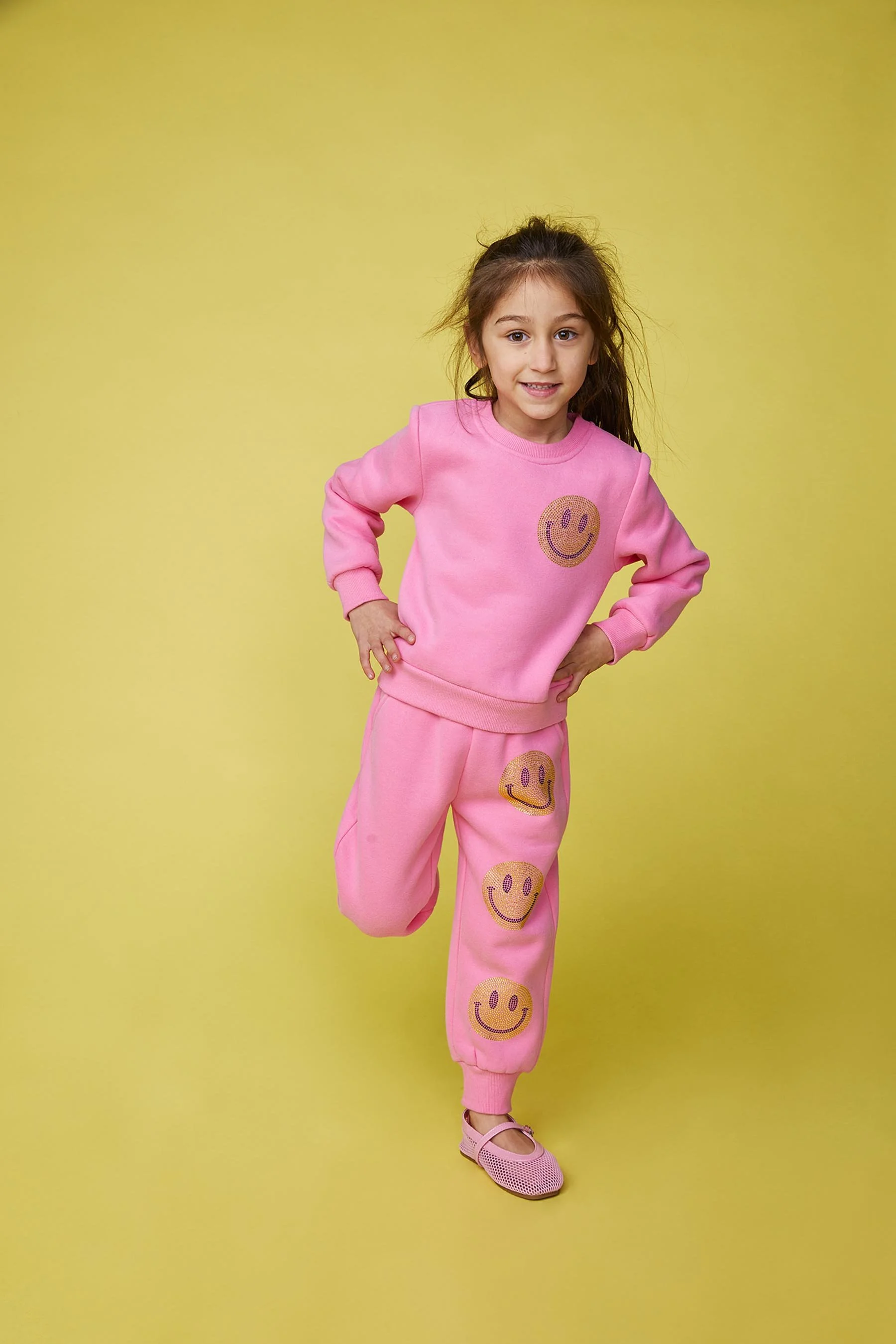 A young girl with long brown hair wearing a pink sweatshirt and matching sweatpants with smiley face designs, standing in front of a yellow background and smiling at the camera.