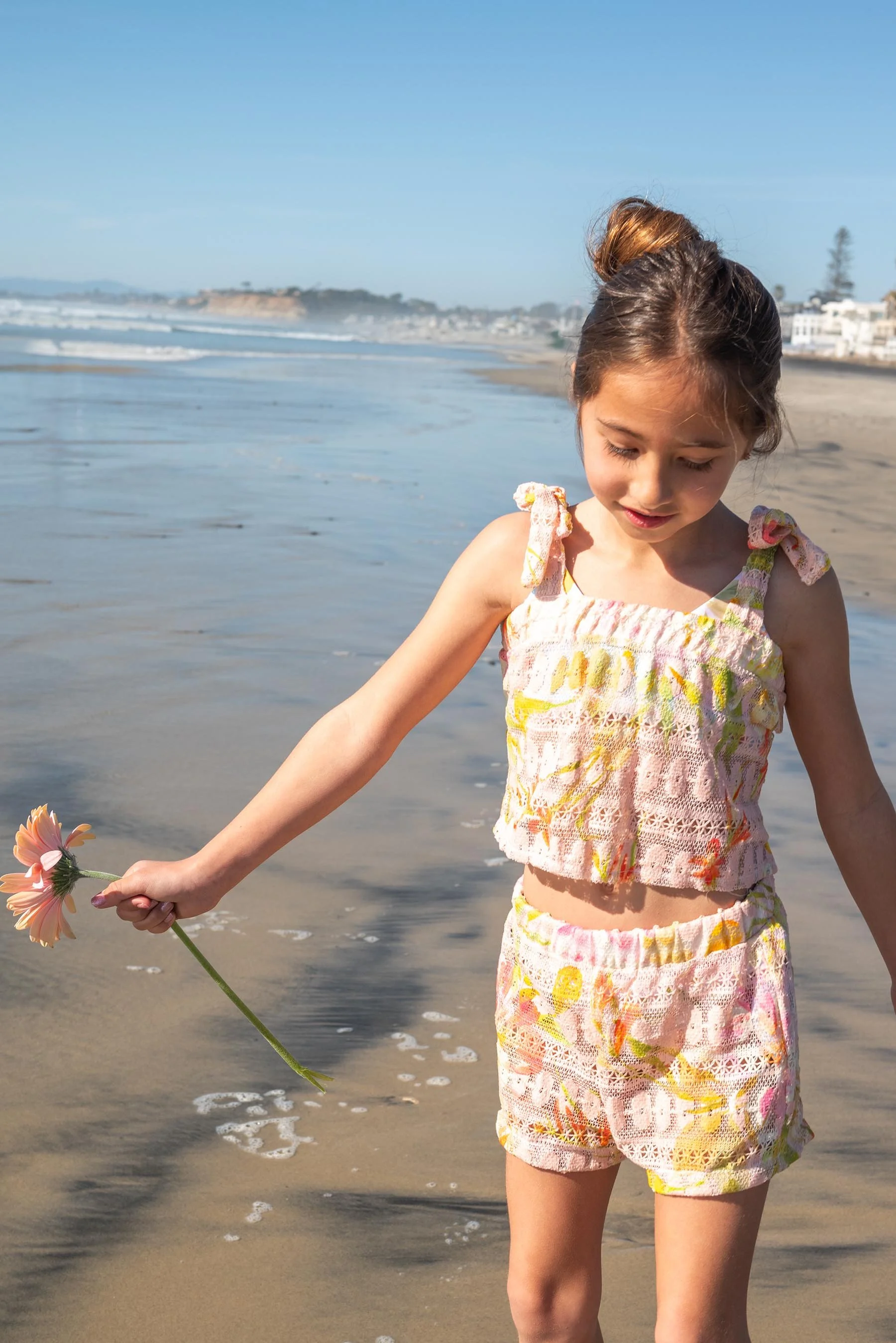 A young girl in a colorful tank top and shorts holding a flower while walking along the beach.