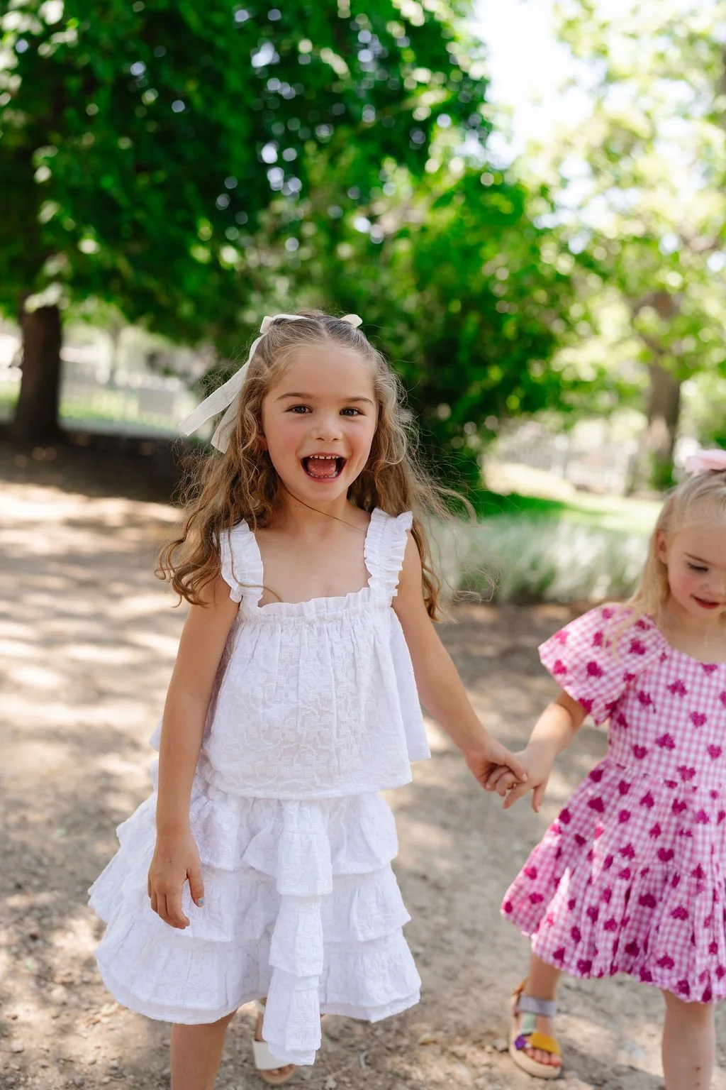 Two young girls holding hands outdoors, one with curly hair wearing a white dress, smiling, and the other with straight hair in a pink checkered dress, partially visible, in a park with green trees and sunlight.