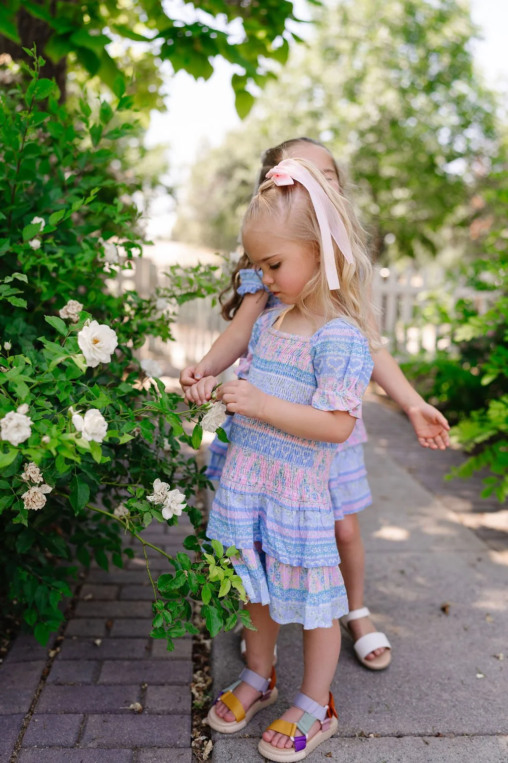 Two young girls in pastel dresses and sandals pick white flowers from a lush green bush on a garden path.