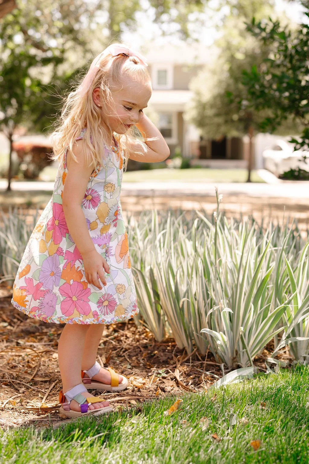 A young girl with long blonde hair wearing a colorful floral dress and sandals stands outdoors near ornamental grass and trees, with a house in the background.