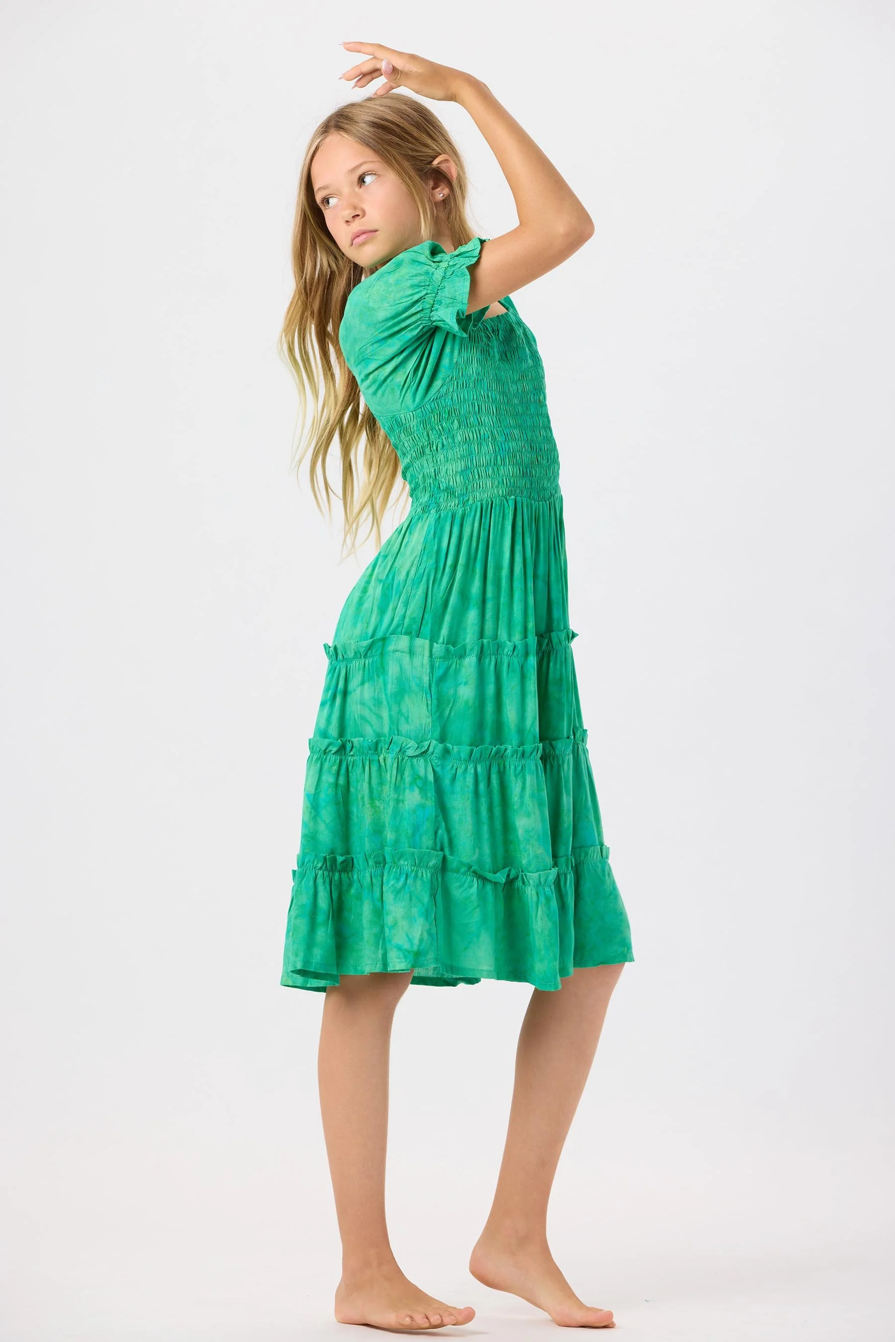 Young girl with long blonde hair wearing a green tiered dress with puff sleeves, standing barefoot against a plain white background, posing with one arm raised above her head.