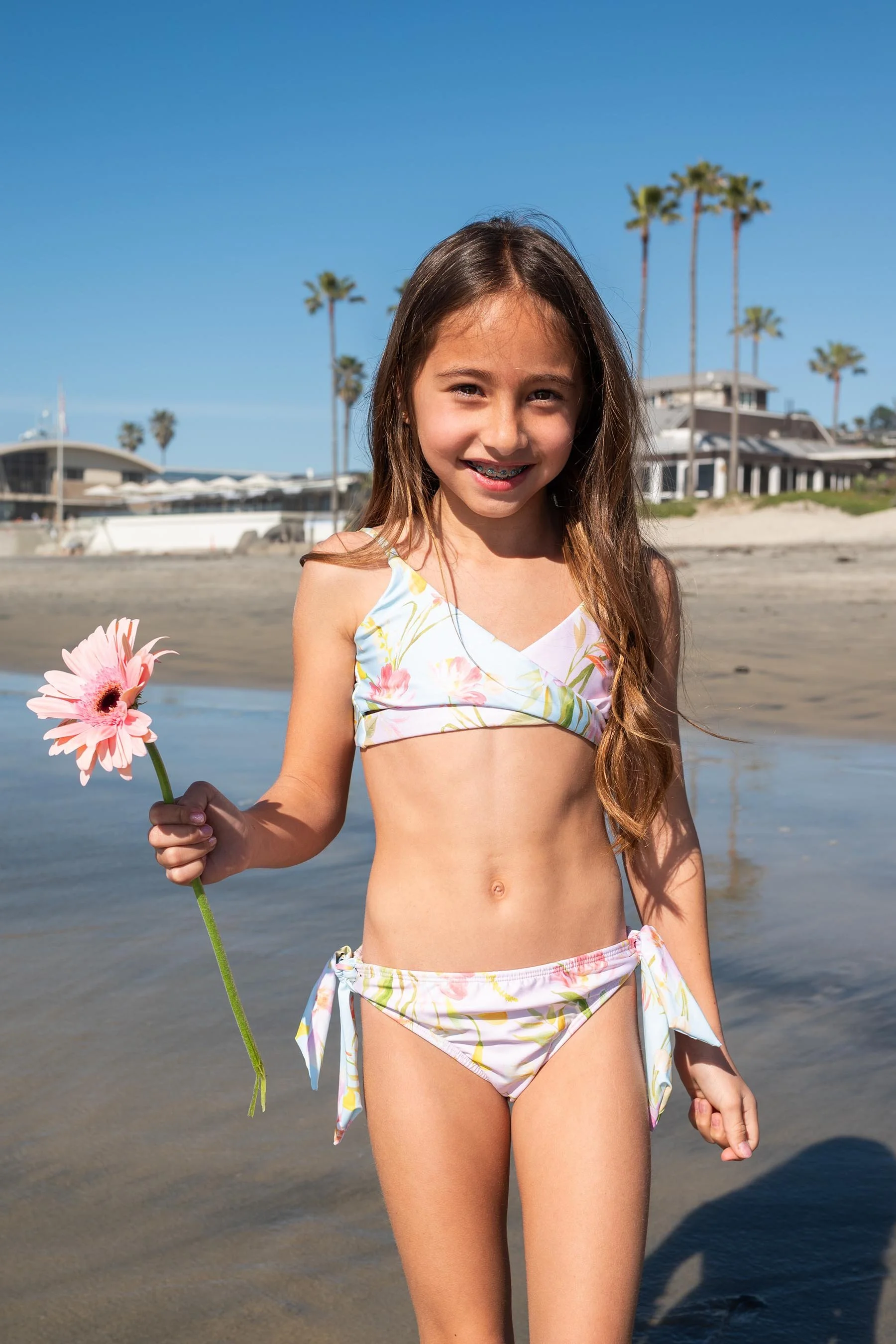 A young girl with long brown hair, braces, wearing a floral bikini, smiling and holding a pink flower, standing on a beach with sand, water, palm trees, and houses in the background on a sunny day.