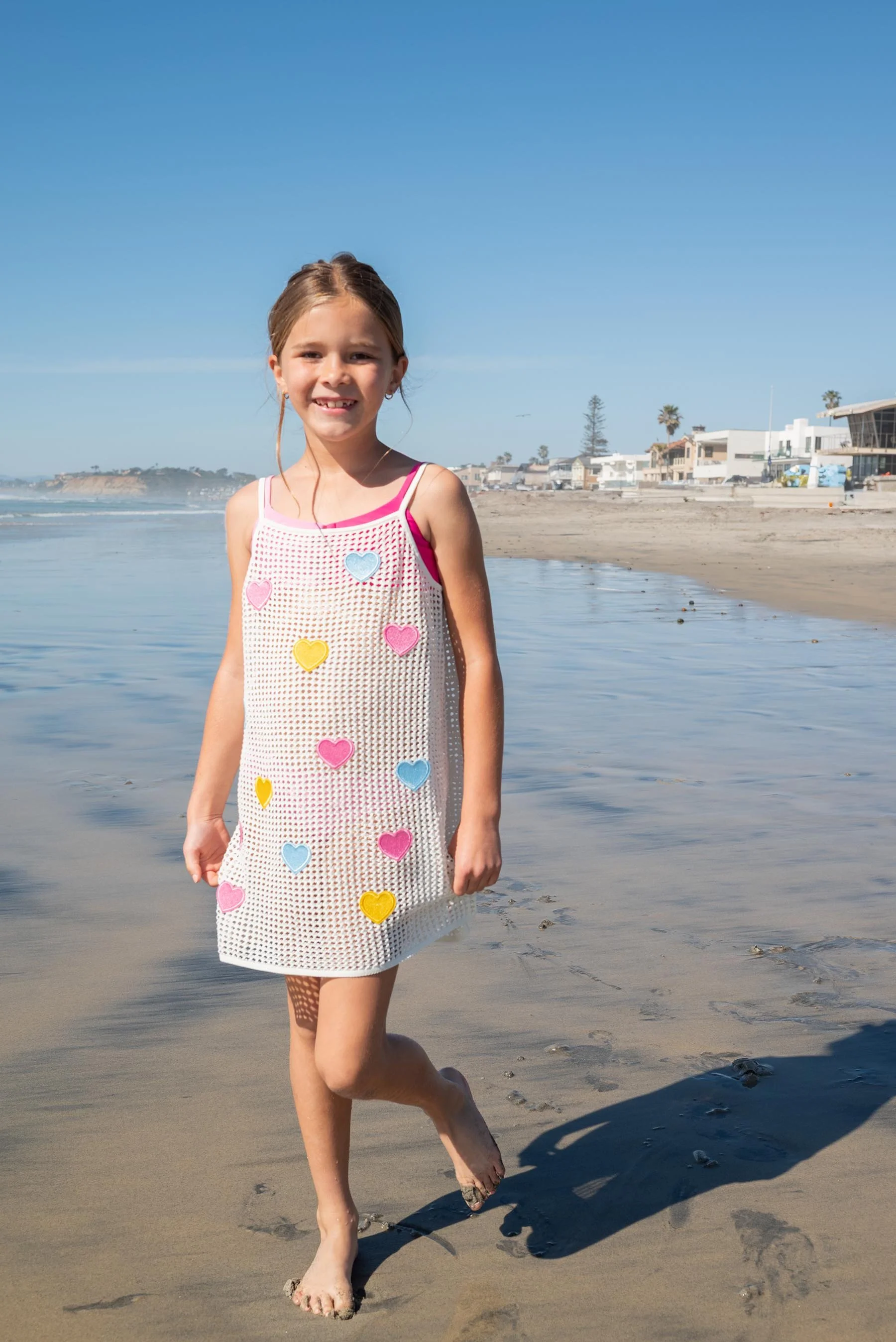A young girl walking barefoot on the beach near the shoreline during daytime, wearing a white mesh dress decorated with colorful heart shapes, smiling at the camera with a background of houses and palm trees.