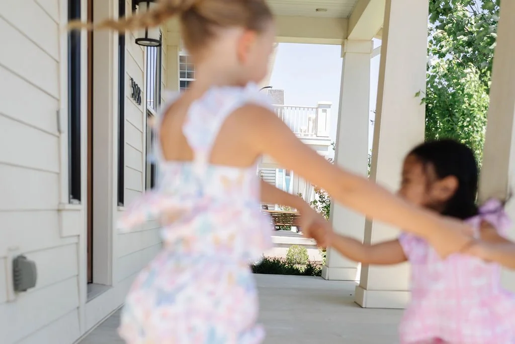 Two young girls in pastel dresses holding hands and playing outside on a porch with trees and houses in the background.