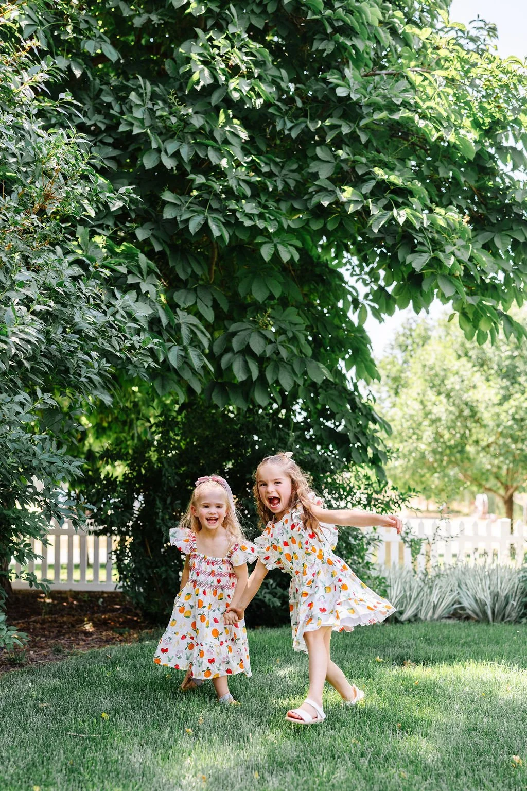Two young girls in floral dresses happily playing together on a grassy lawn under a large leafy tree, holding hands and smiling.