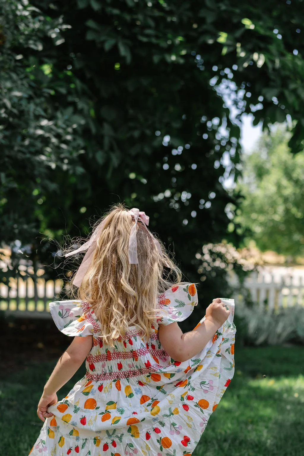 A young girl with long, curly blonde hair, wearing a colorful dress with fruit and floral patterns, runs through a lush green backyard with a white picket fence and large trees.
