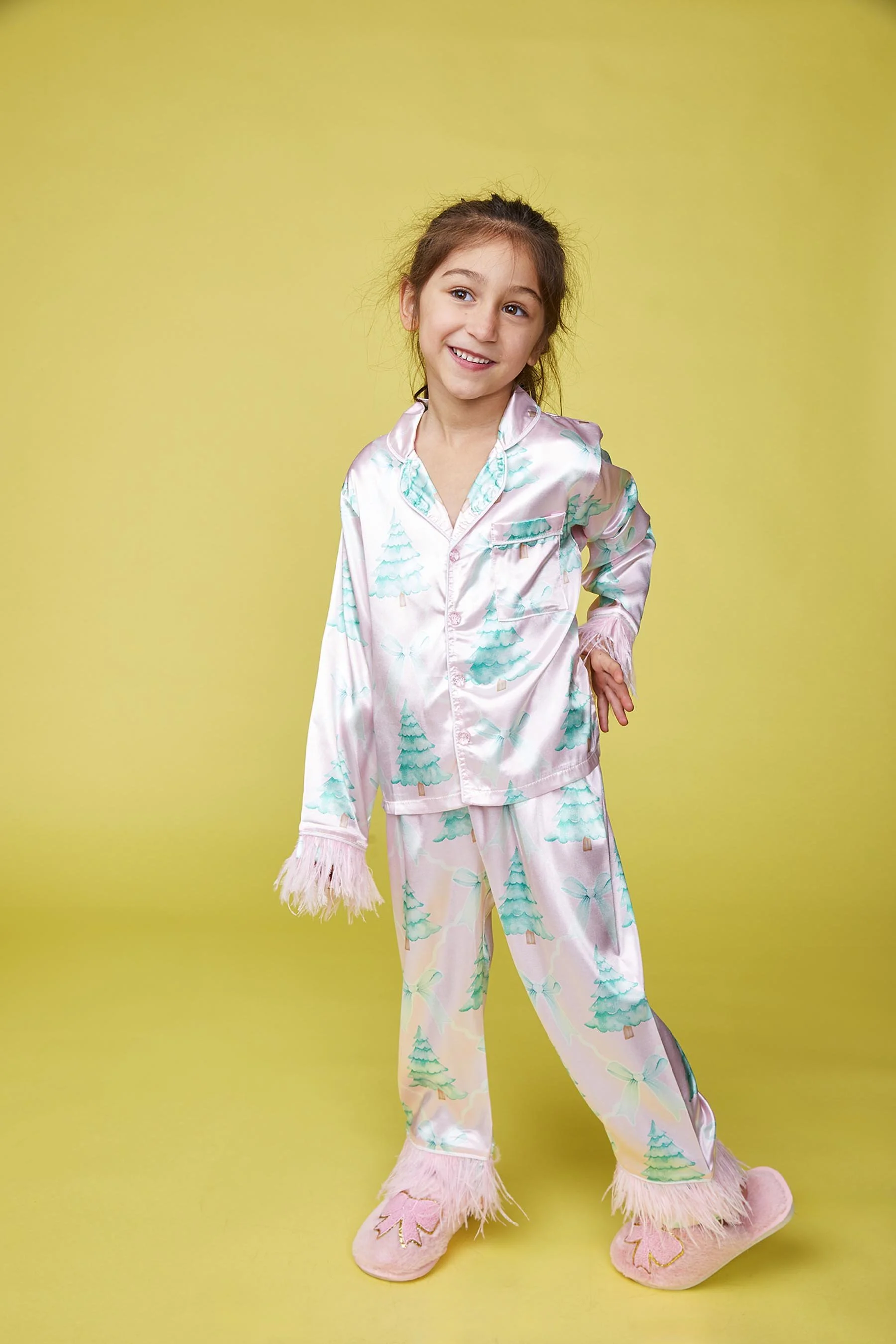 Young girl in pastel Christmas pajamas with trees and bows, standing on a yellow background.