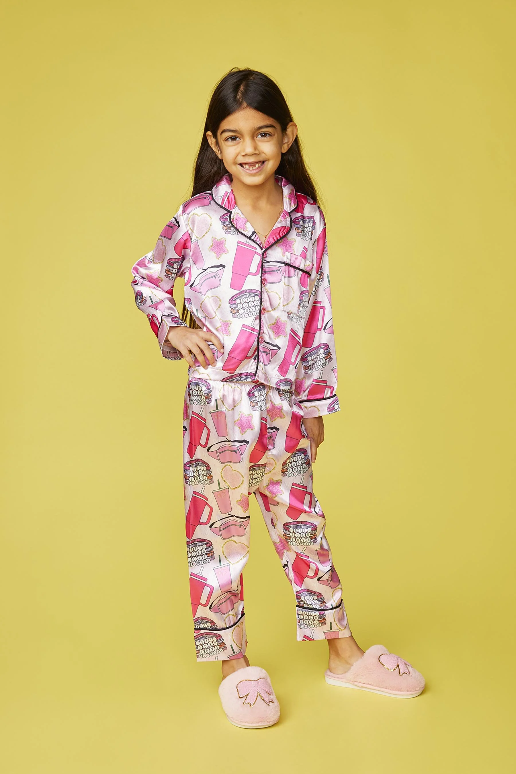 A young girl in pink and white pajamas with makeup and lipstick patterns standing against a yellow background, smiling, with one hand on her hip and wearing pink slippers.