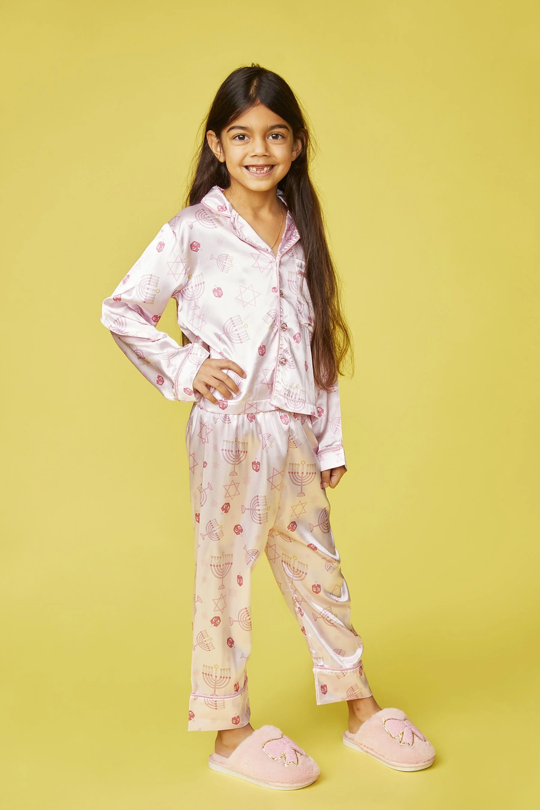 A young girl wearing pink pajamas and pink slippers, standing confidently with a smile against a yellow background.