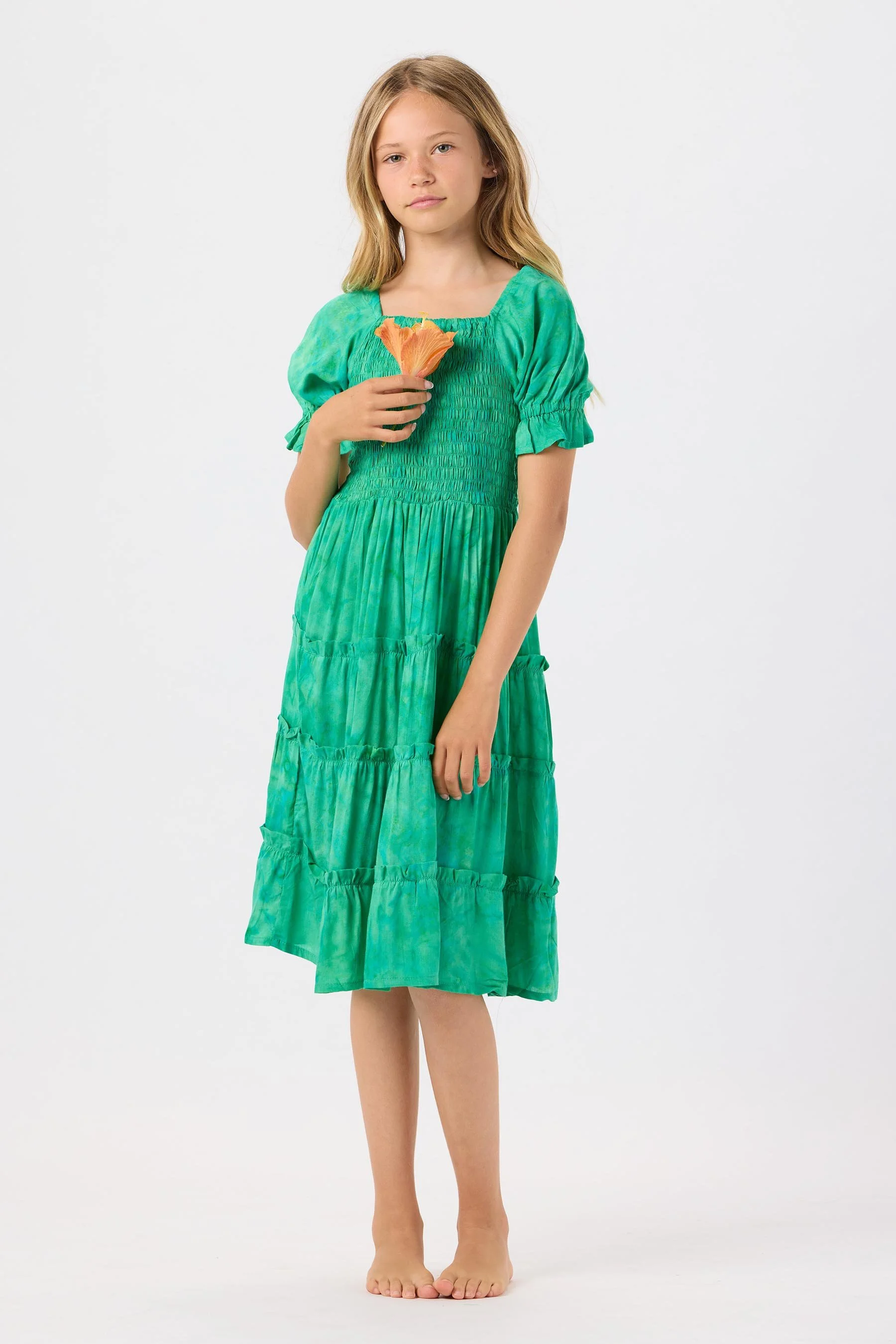 A young girl in a green, tie-dye dress standing barefoot, holding an orange flower near her chest, with a neutral expression against a plain white background.