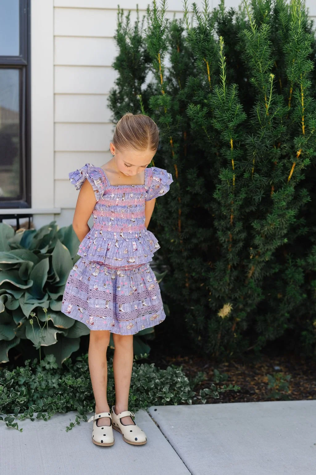 A young girl in a purple dress with ruffles and a pattern, standing outdoors on a sidewalk with her head bowed and hands behind her back, near green shrubs and plants.