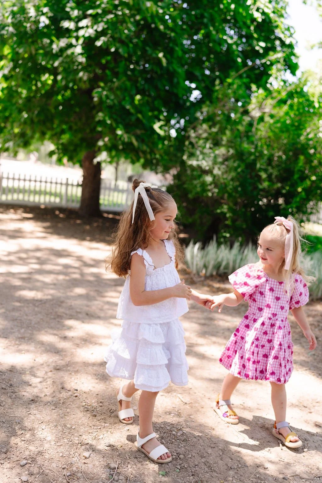 Two young girls holding hands and playing outdoors on a sunny day with green trees in the background.