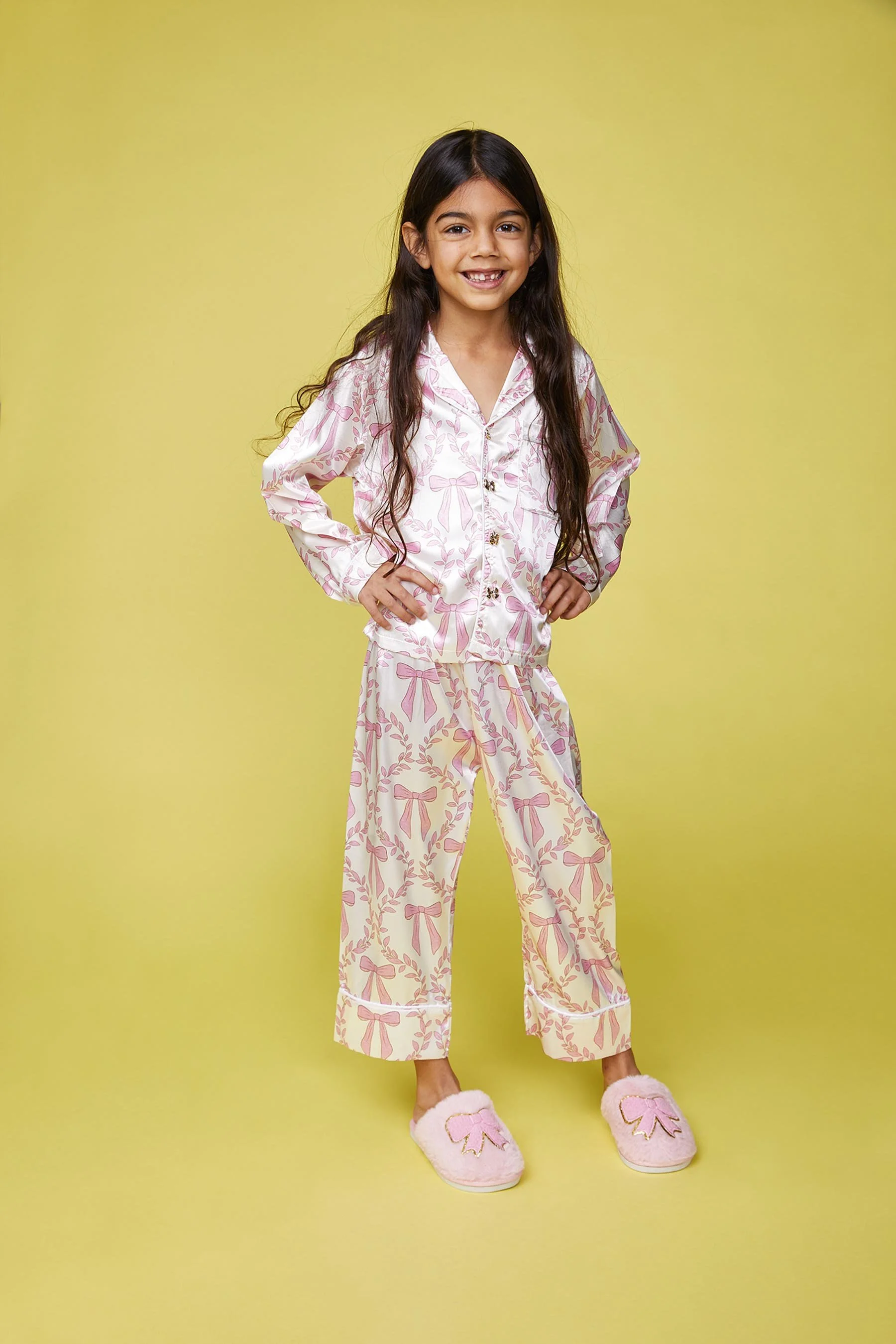 A young girl with long dark hair wearing pink and white pajama set with bow and wreath patterns, standing against a yellow background, smiling with hands on hips and wearing fuzzy pink slippers with bow designs.