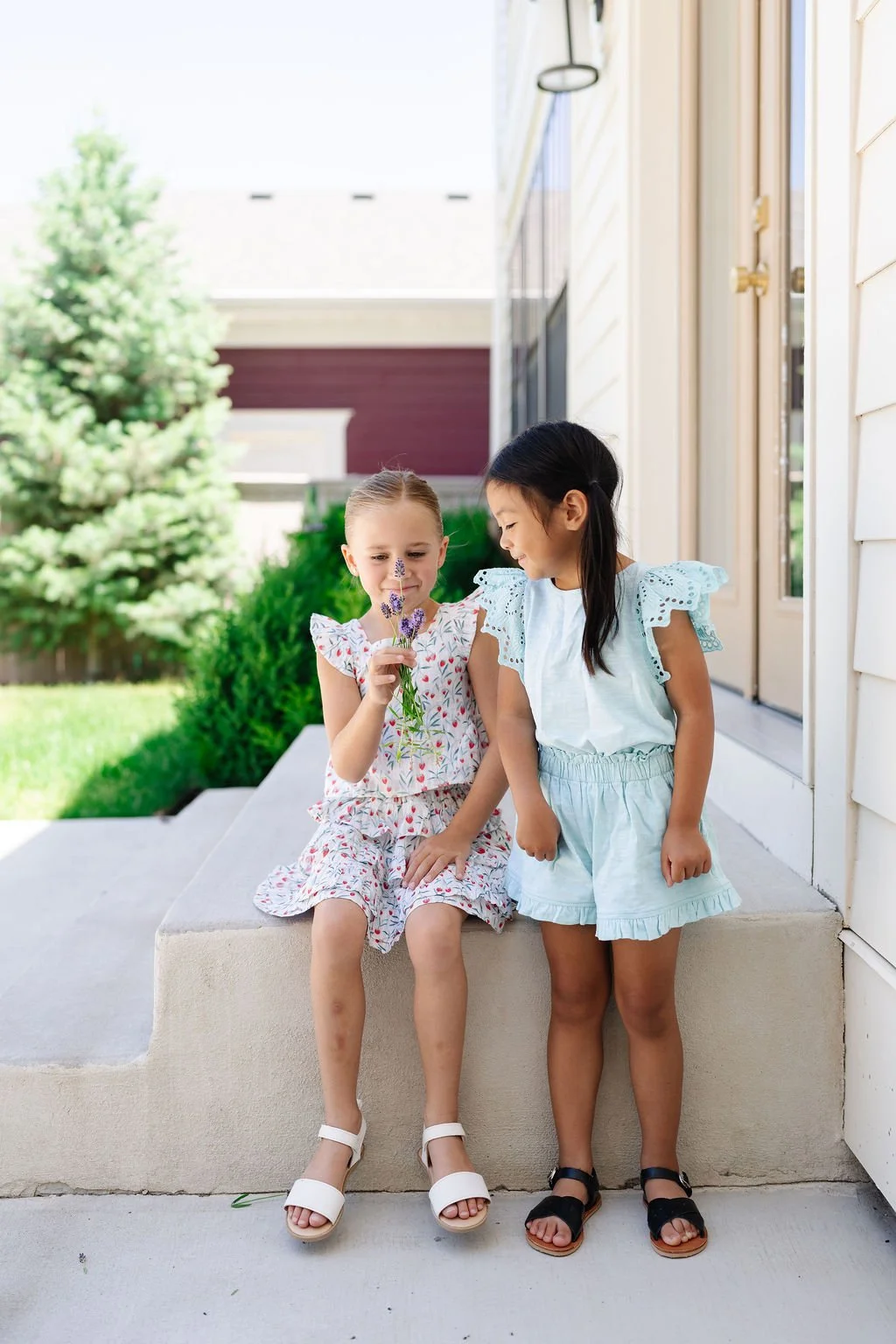 Two young girls sitting on a porch step, one girl is holding flowers and smiling at the other girl, who is smiling back.