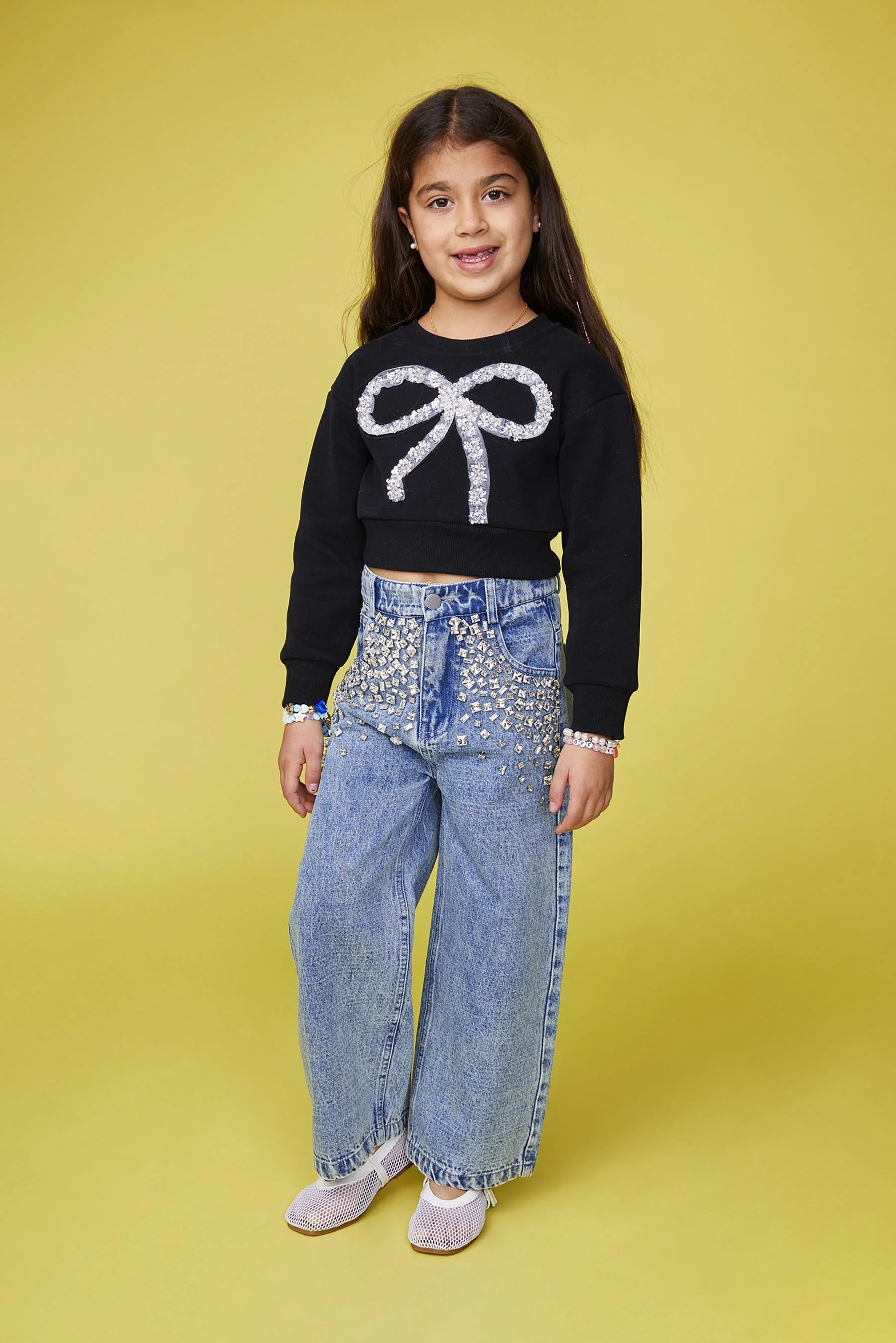 Young girl wearing a black cropped sweater with a sparkling bow design, high-waisted baggy jeans with silver embellishments, white shoes, and colorful bracelets, standing against a yellow background.