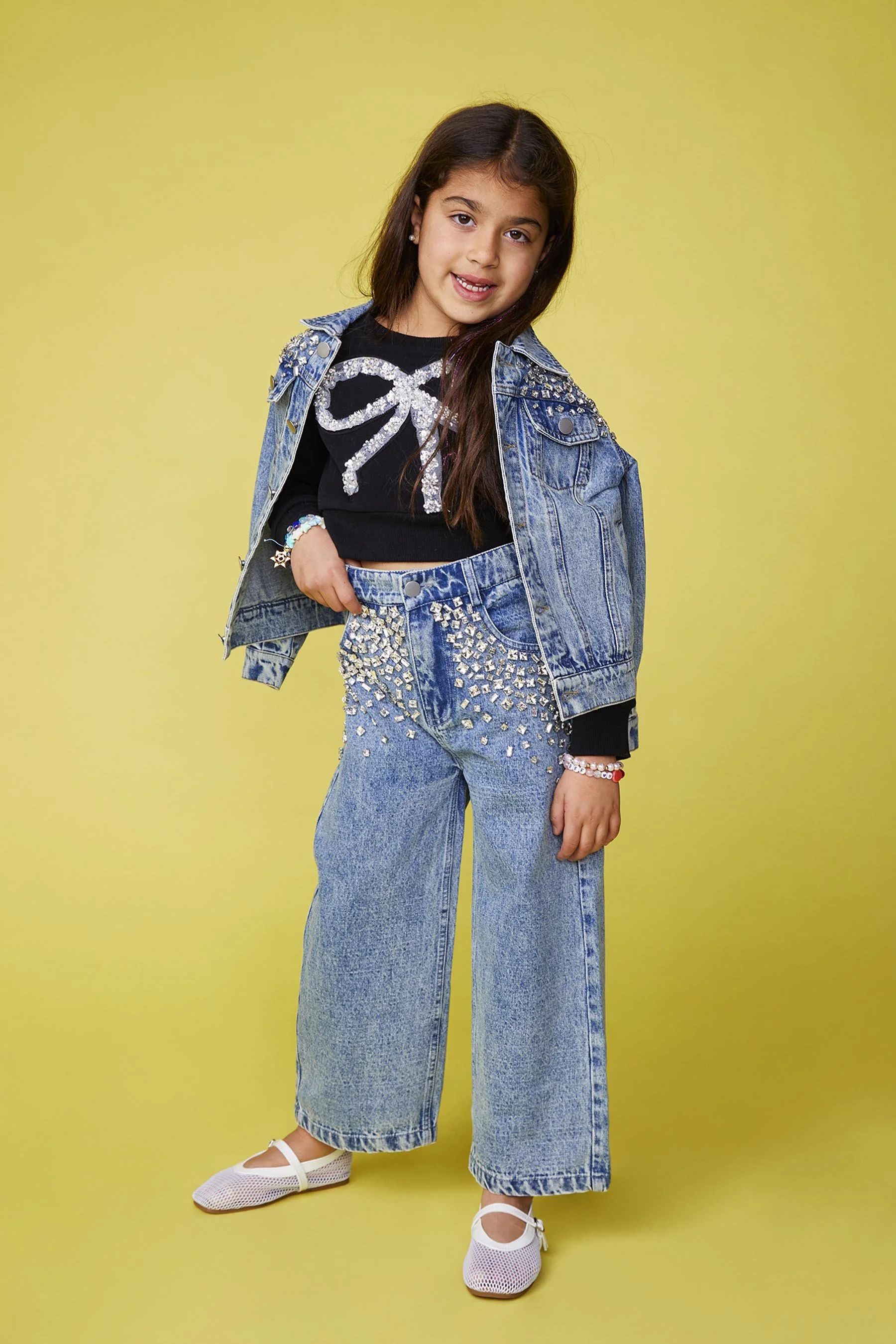A young girl with long dark hair wearing a black top with a bow design, a denim jacket, wide-leg jeans embellished with sparkly accents, white shoes, and jewelry, against a yellow background.