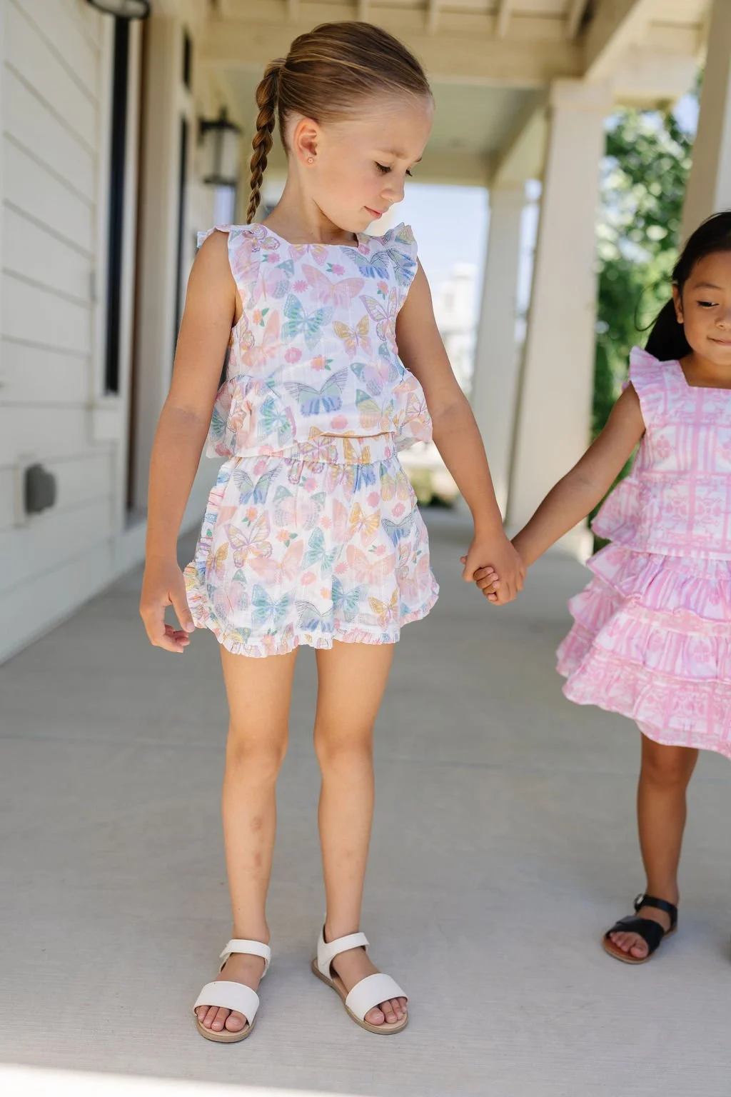 Two young girls holding hands on a porch, wearing matching pastel-colored butterfly and pink patterns dresses, with one girl wearing white sandals and the other in black sandals.