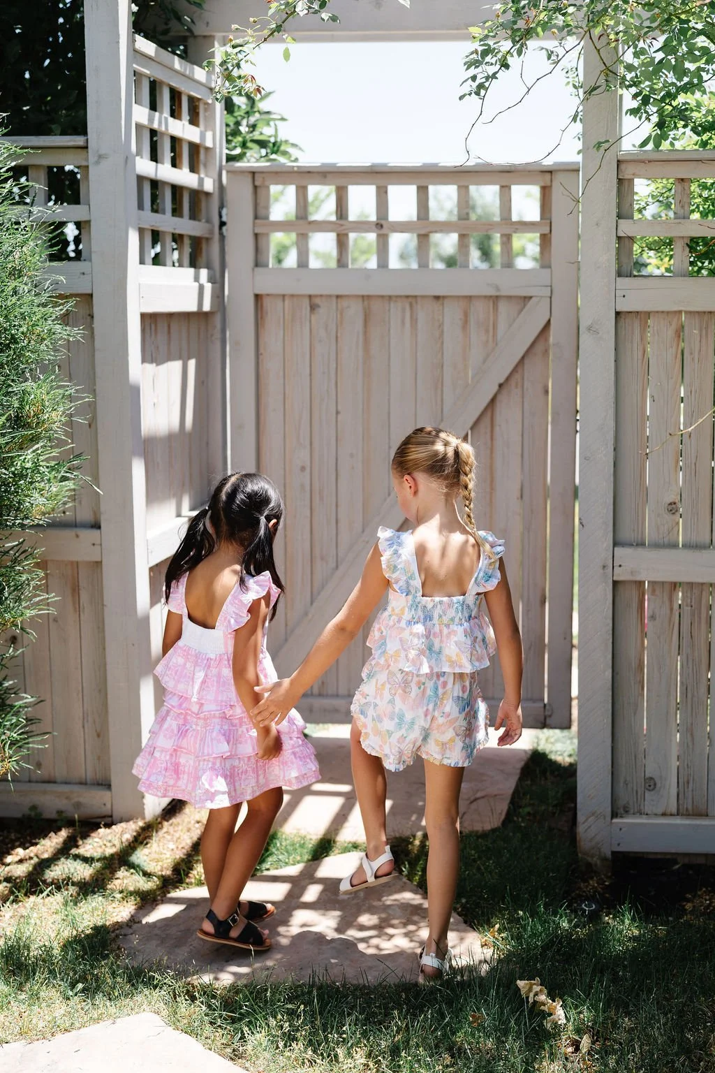 Two young girls in pastel-colored dresses holding hands and walking through a wooden garden gate on a sunny day.