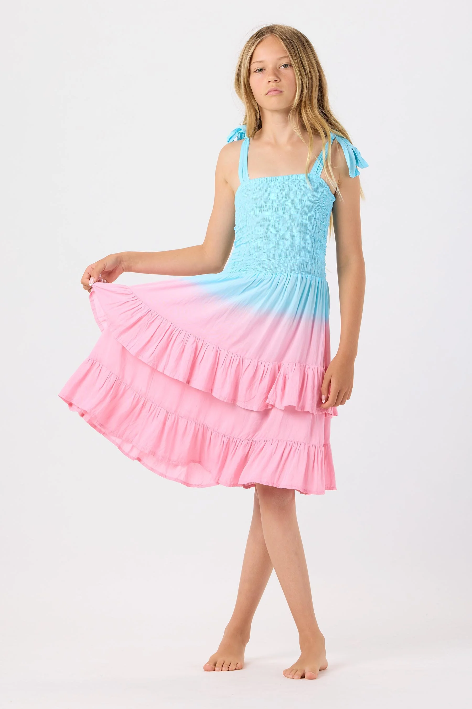 Young girl in a sleeveless, multicolored dress with blue and pink ruffles, standing barefoot against a plain white background, holding the skirt out to the side.